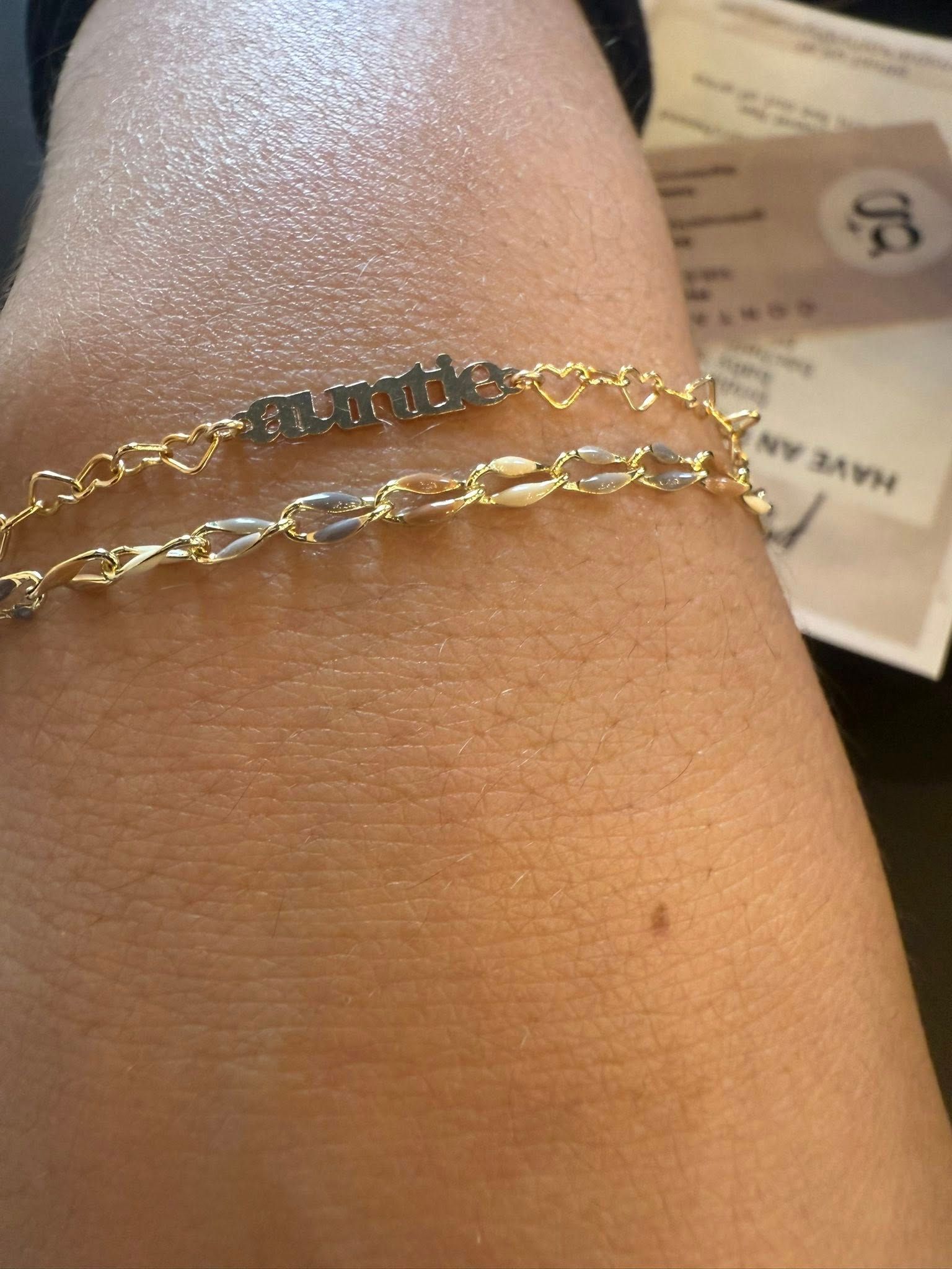 Two gold chain bracelets on a wrist, one with clear stones, against a blurred background.