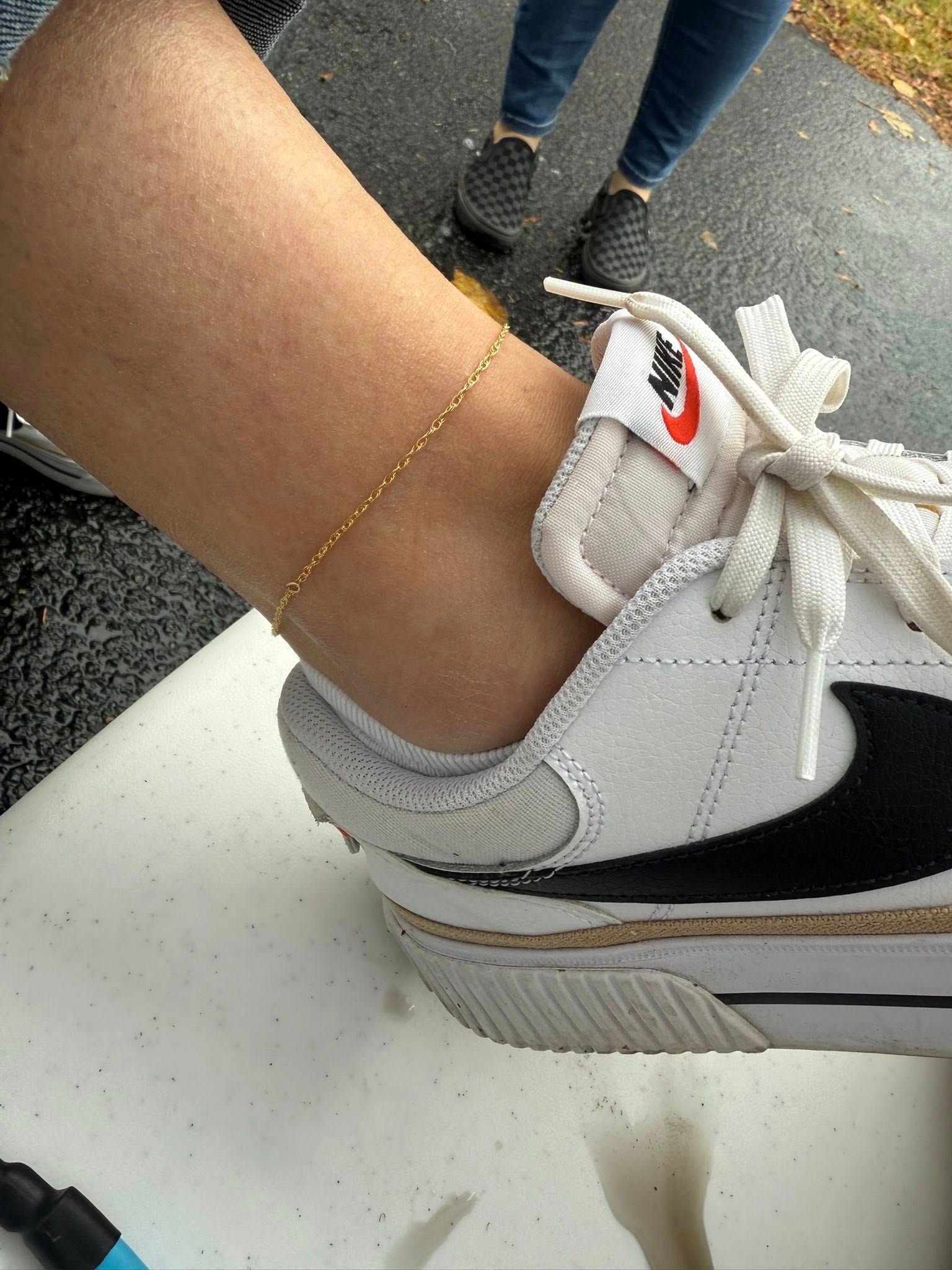 White sneaker on a foot with a thin gold anklet, resting on a white surface.
