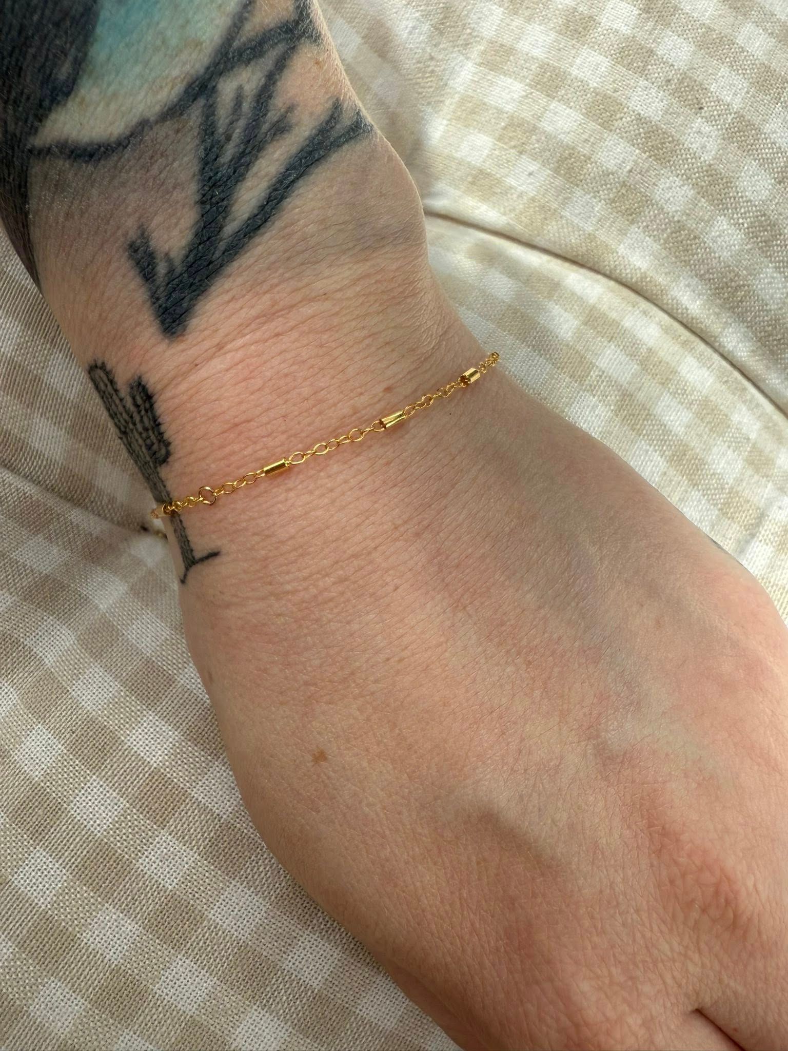 Close-up of a wrist with a small black tattoo and a thin gold bracelet on a beige fabric background