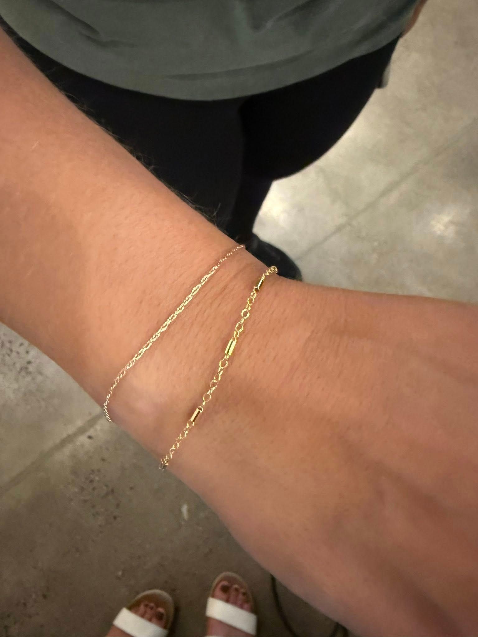 Close-up of a wrist wearing two thin gold bracelets over a tiled floor.