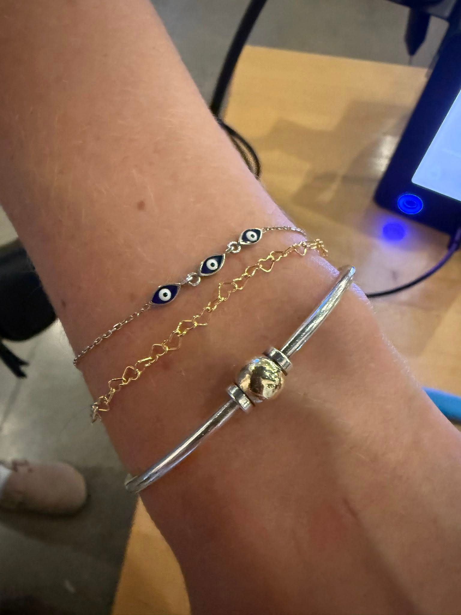 Wrist with three bracelets, including gold and silver chains, on a wooden table nearby