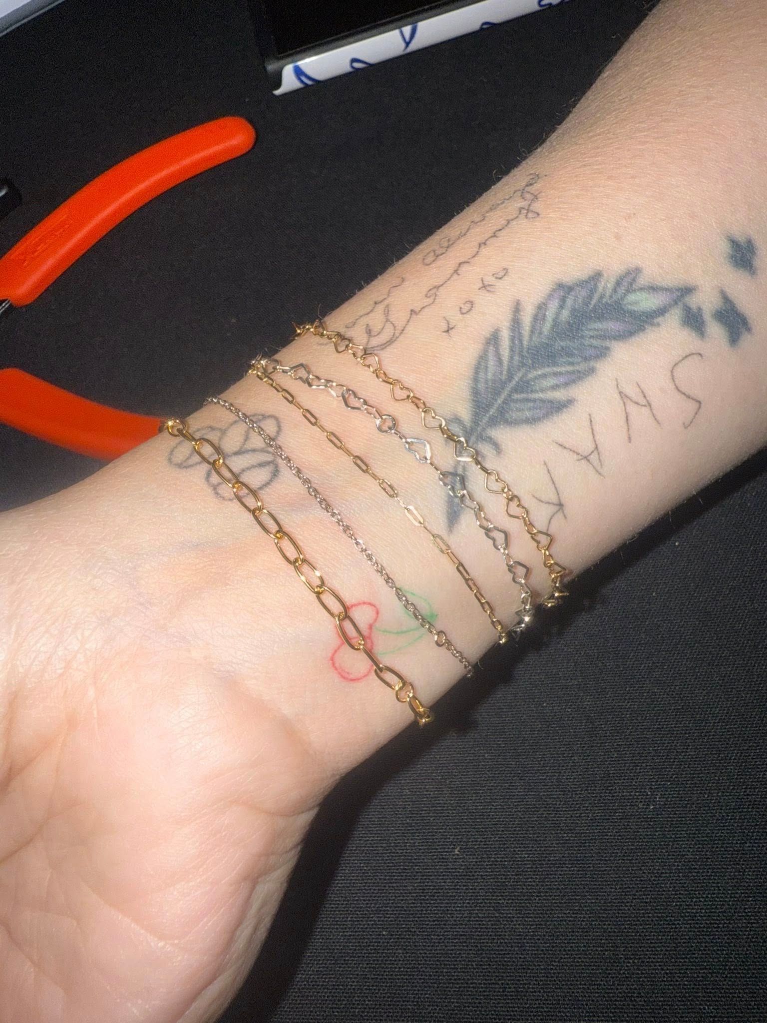 Forearm with feather tattoo and layered gold bracelets on a dark fabric background