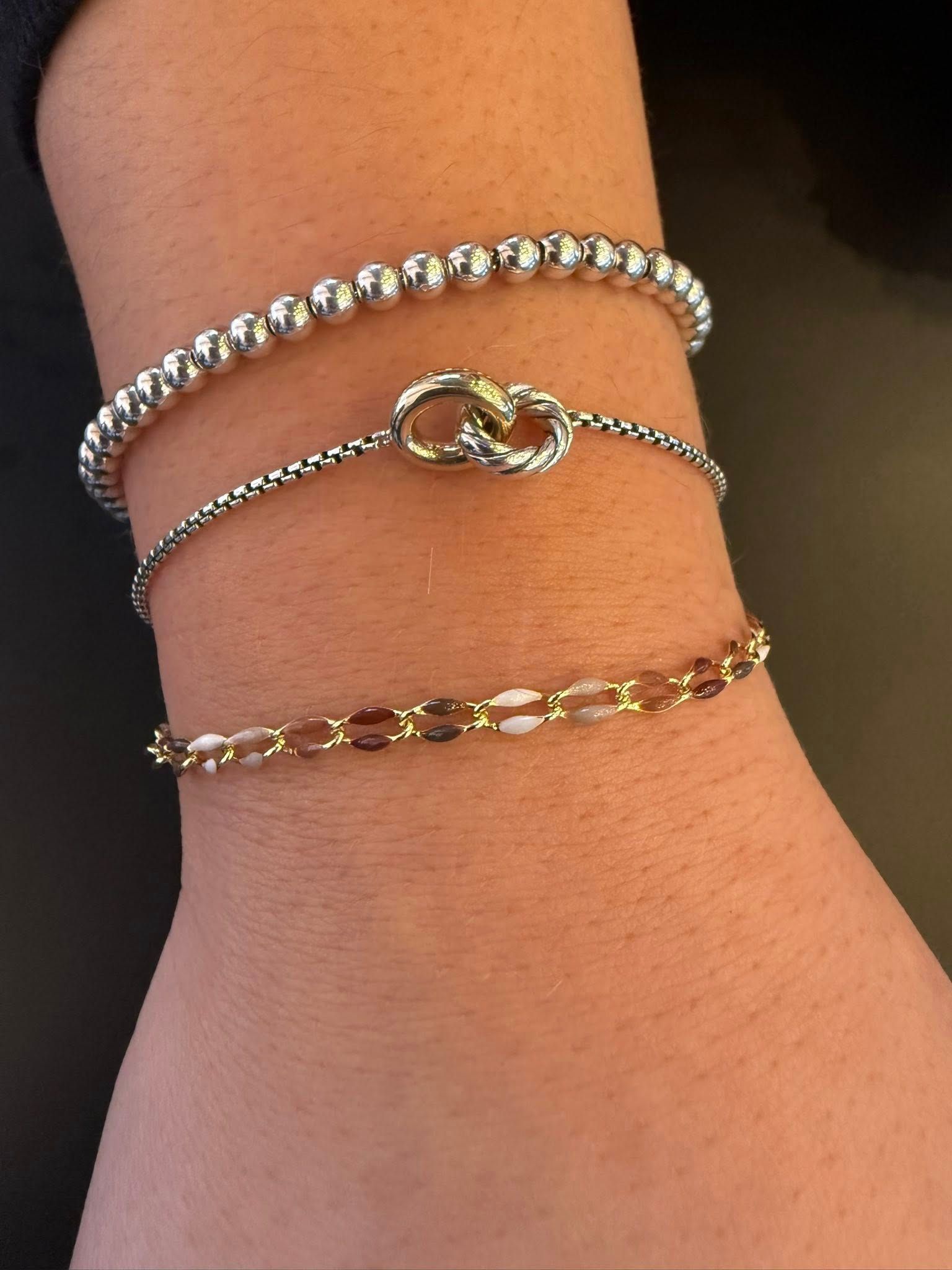 Three layered bracelets on a wrist: silver chain, gold knot, and gold link band.