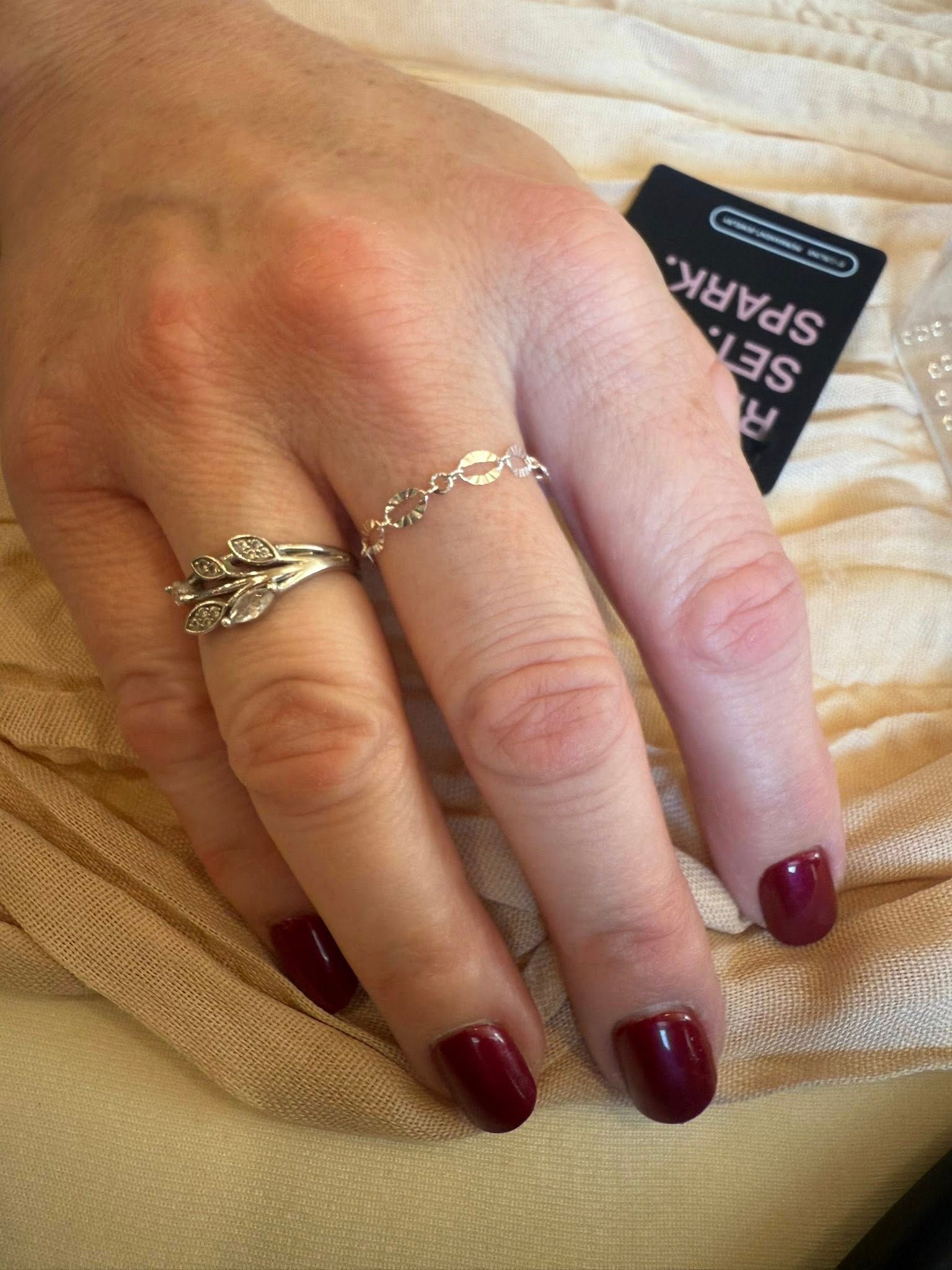 Hand with burgundy nails wearing two delicate gold rings on a beige knitted fabric.