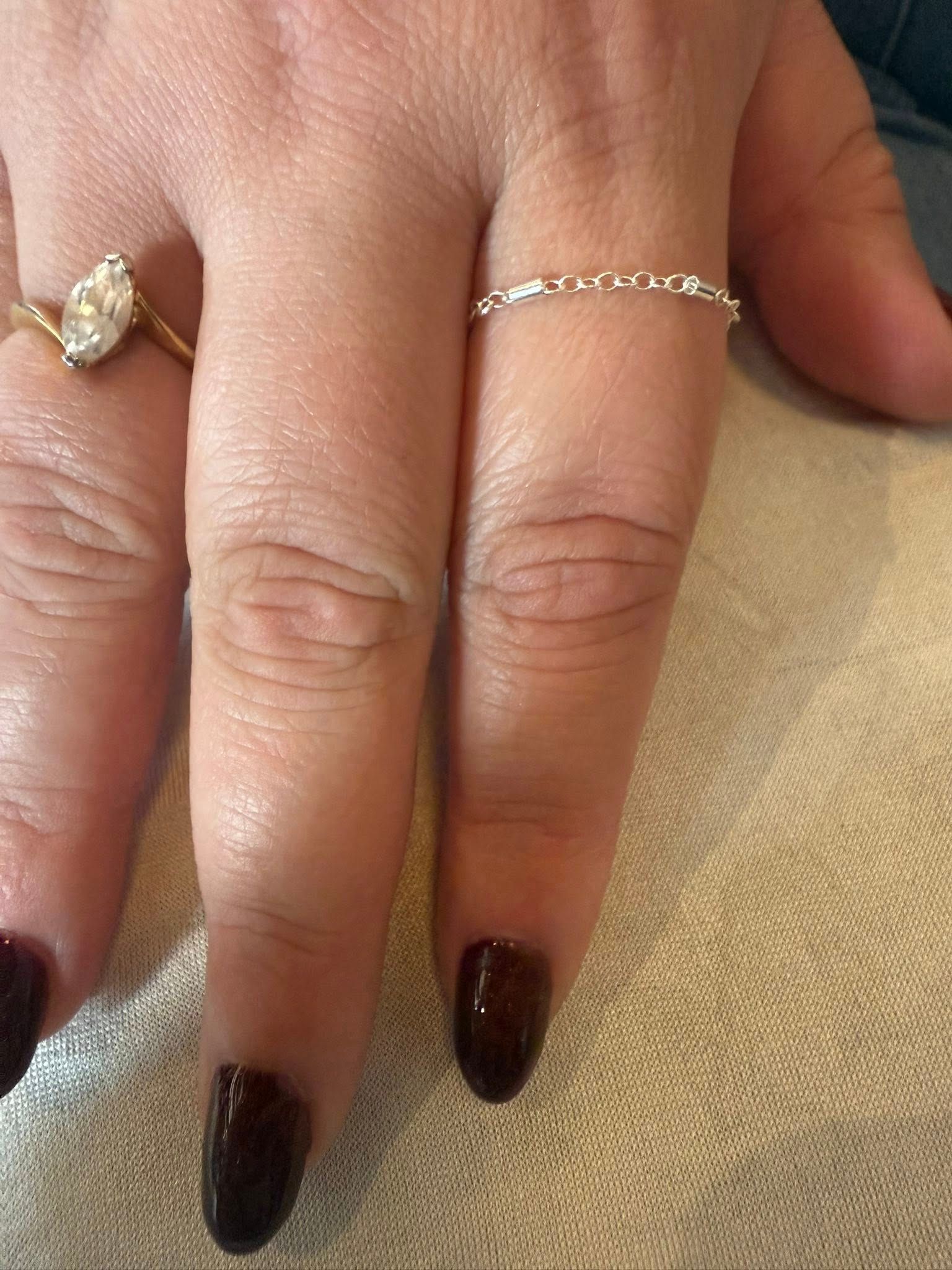 Hand with dark burgundy manicured nails and thin gold rings resting on beige fabric.