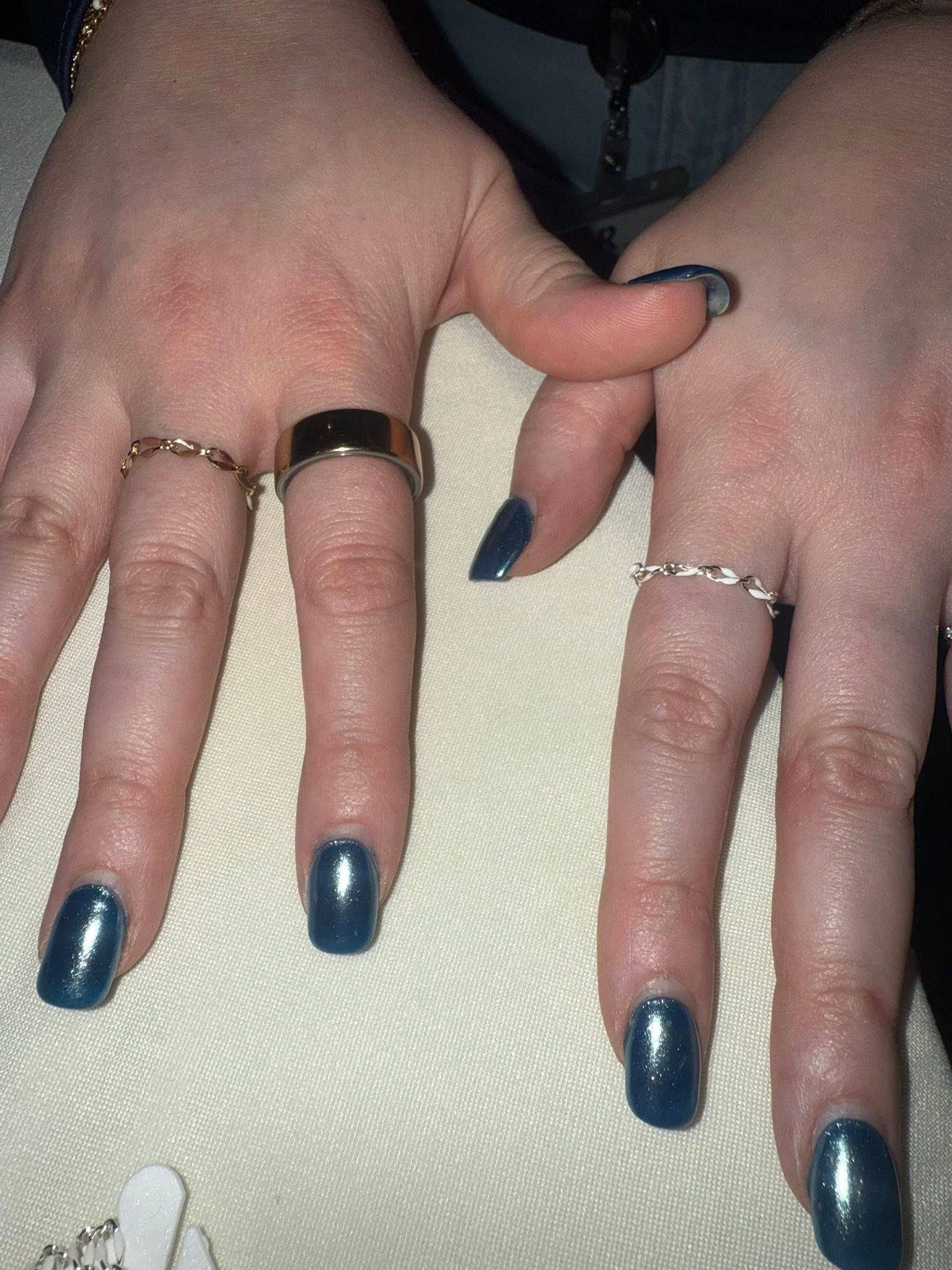 Hands with dark blue manicured nails and several rings resting on a light fabric surface.