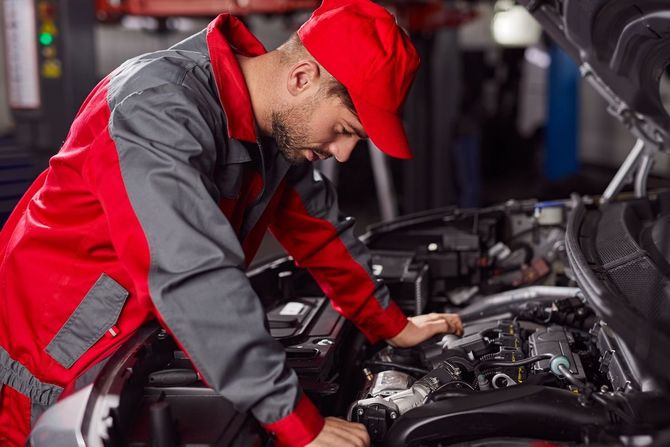 Mechanic in red and gray uniform inspecting a car engine in a garage