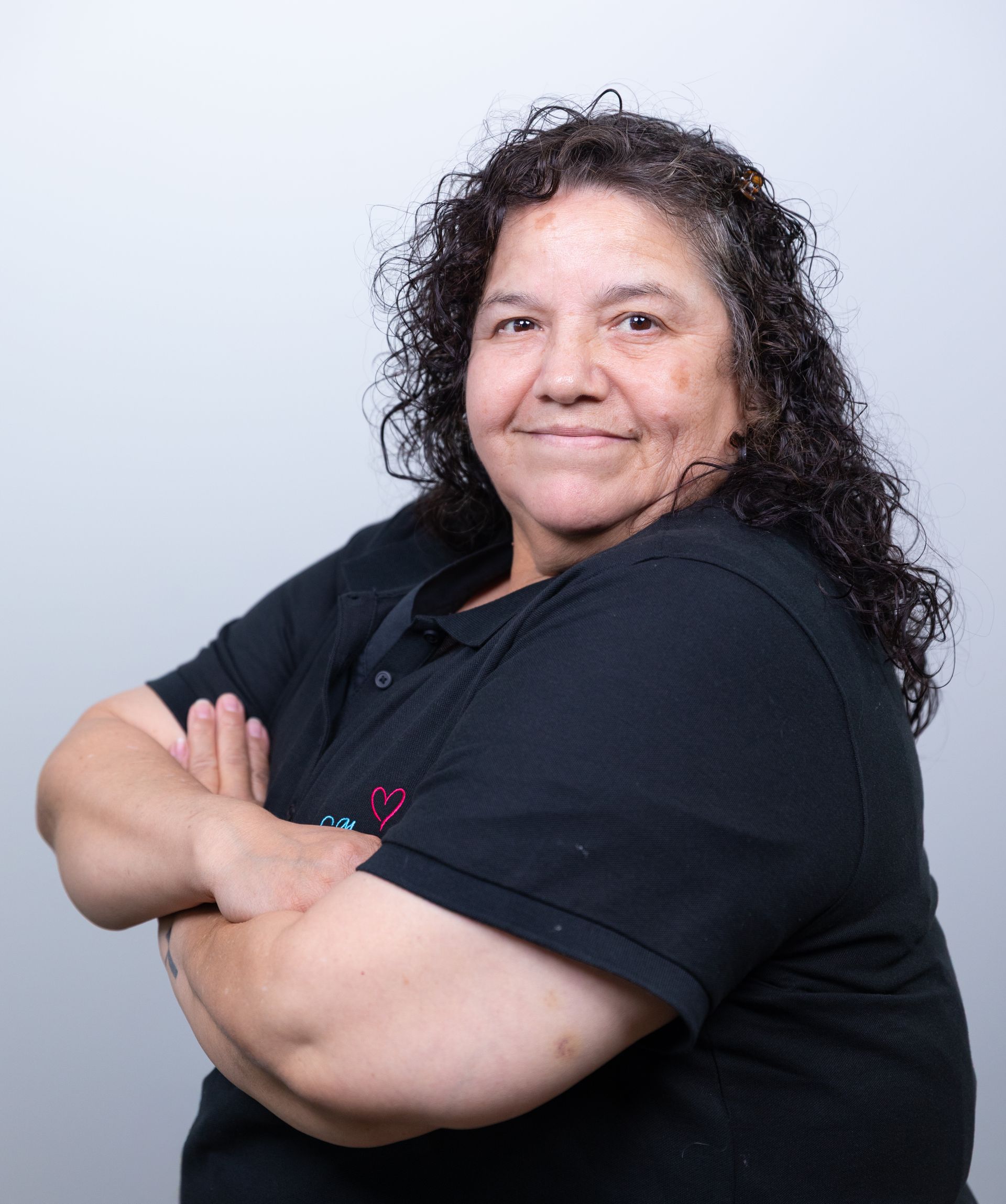 Woman with curly dark hair, arms crossed, wearing a black shirt, smiling against a light gray backdrop.