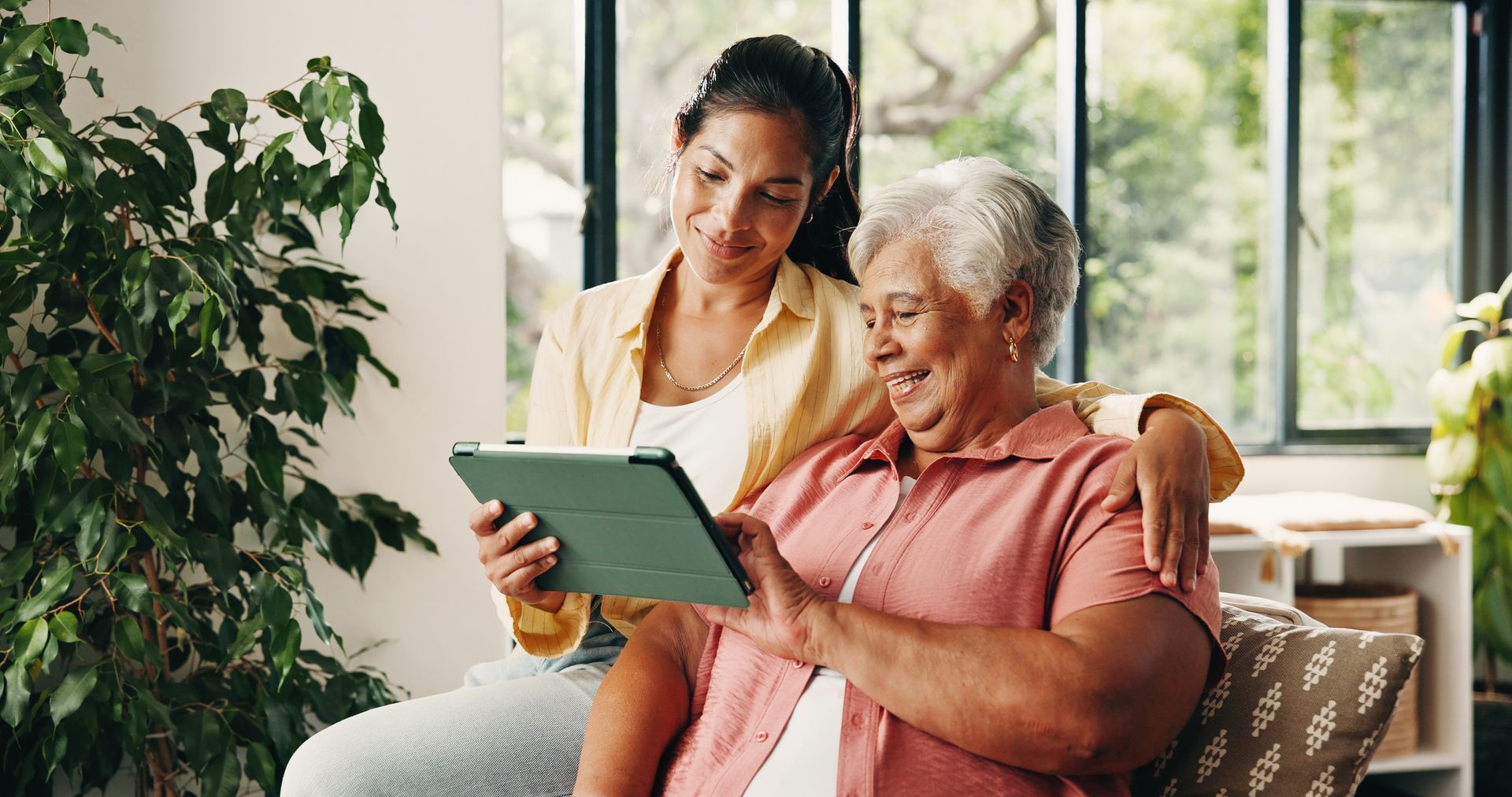 Woman and older adult sharing a tablet on a sofa in a bright living room
