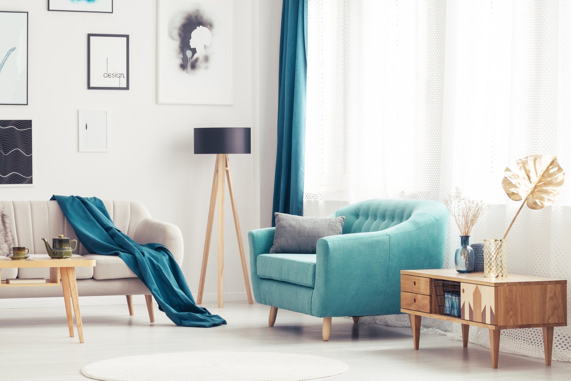 White loungroom with multiple wall hangings and opaque sheets, framed and accented by turquoise drape, lounge chair and throwrug over neutral couch. Timber light stand, table and dresser as complement.