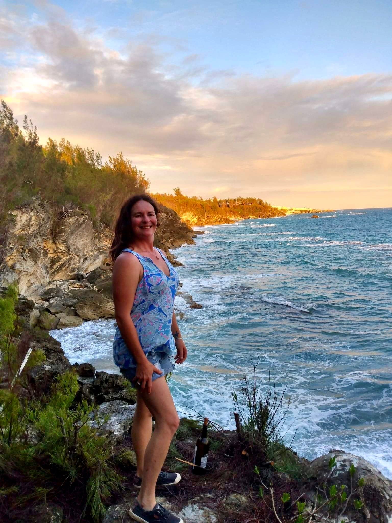 Woman standing on a rocky coastline at sunset, with waves and a glowing sky in the background.