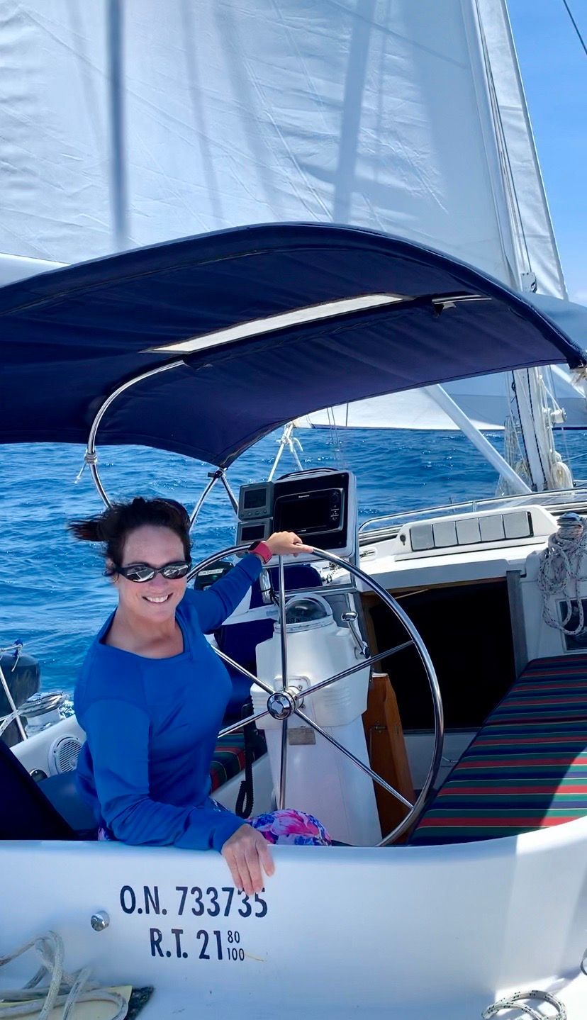 Person smiling at a sailboat’s helm under a blue canopy on a sunny day at sea