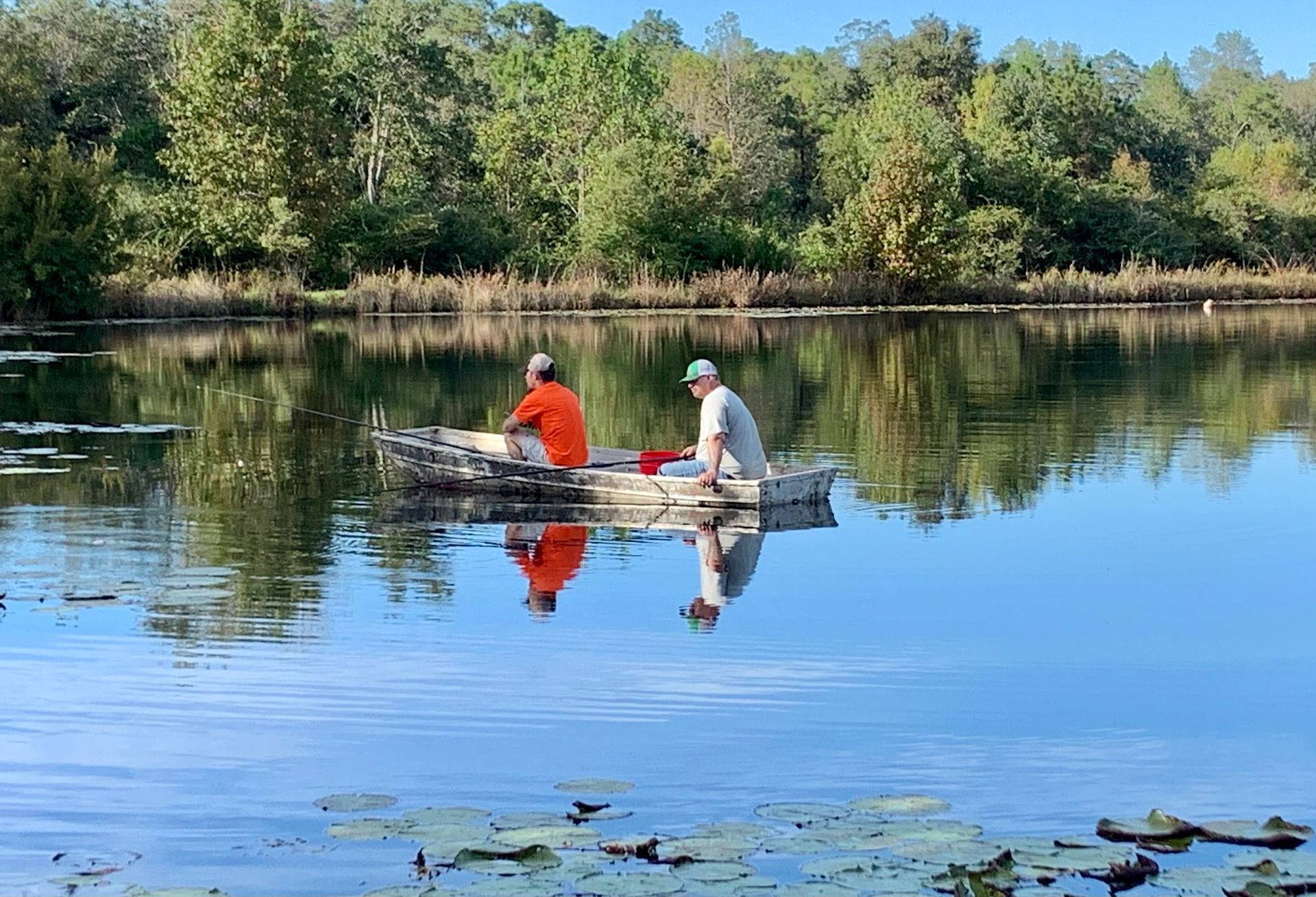 Two people in a small boat on a calm lake, surrounded by trees and reflected in the water.