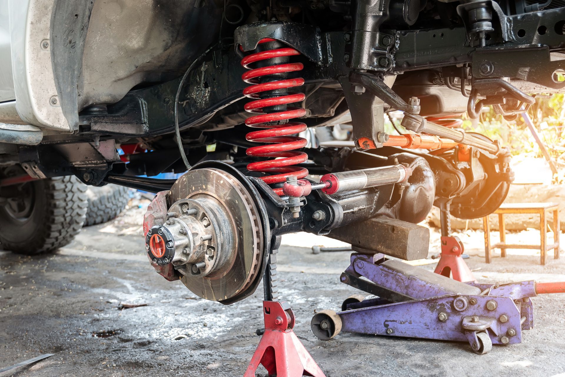 Car's suspension exposed; red coil spring, brake rotor, jack stands, and floor. Repair setting, outdoor.
