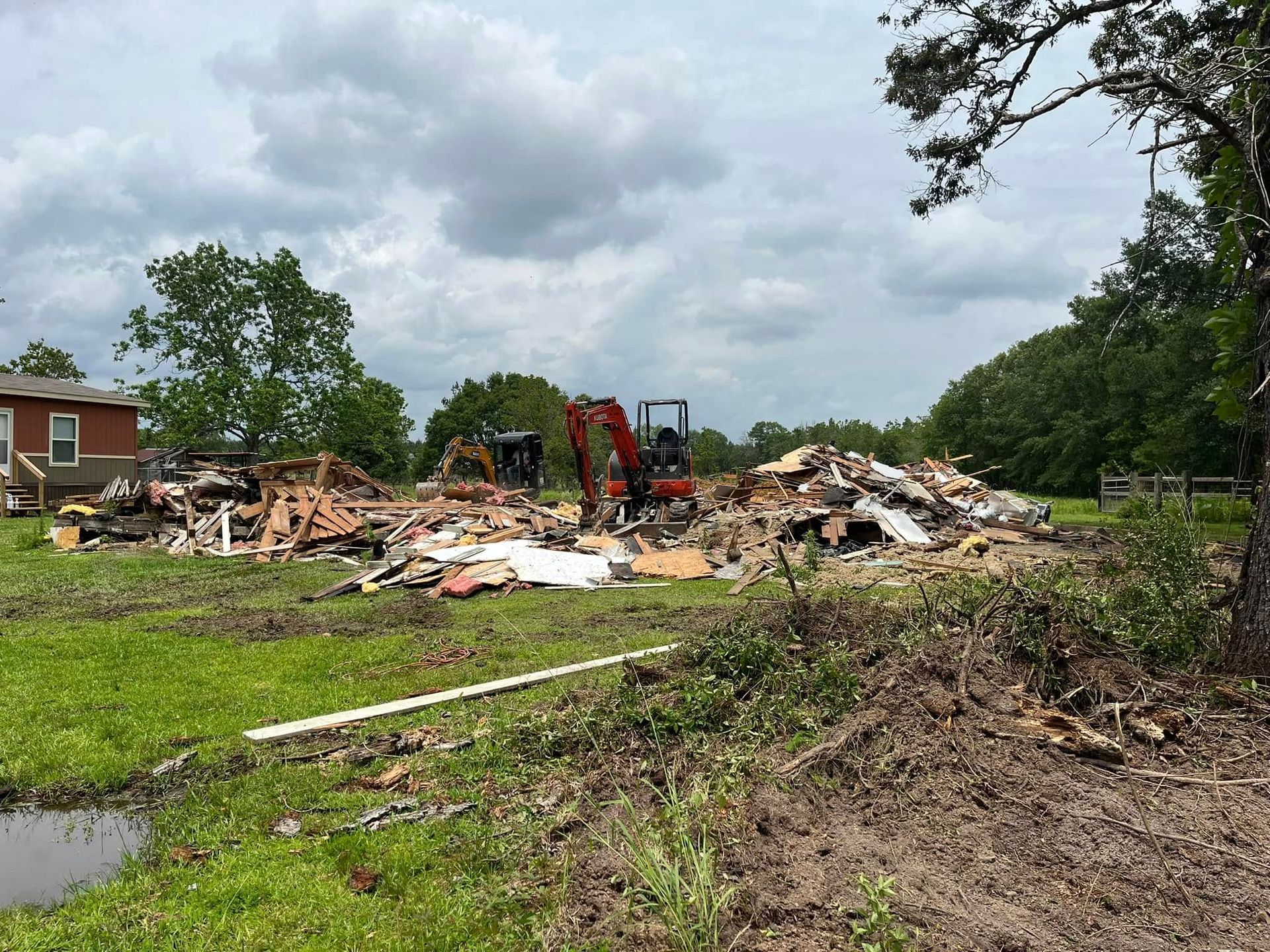 Demolition site with rubble piles, excavator, and cloudy sky beside a grassy lot