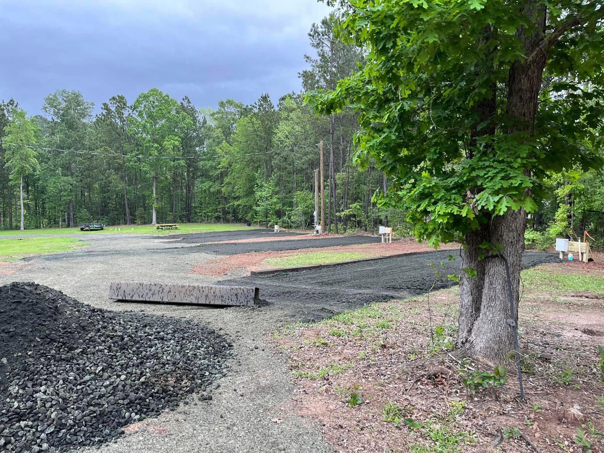 Gravel road under construction beside trees, with dirt mounds and a cleared wooded area.