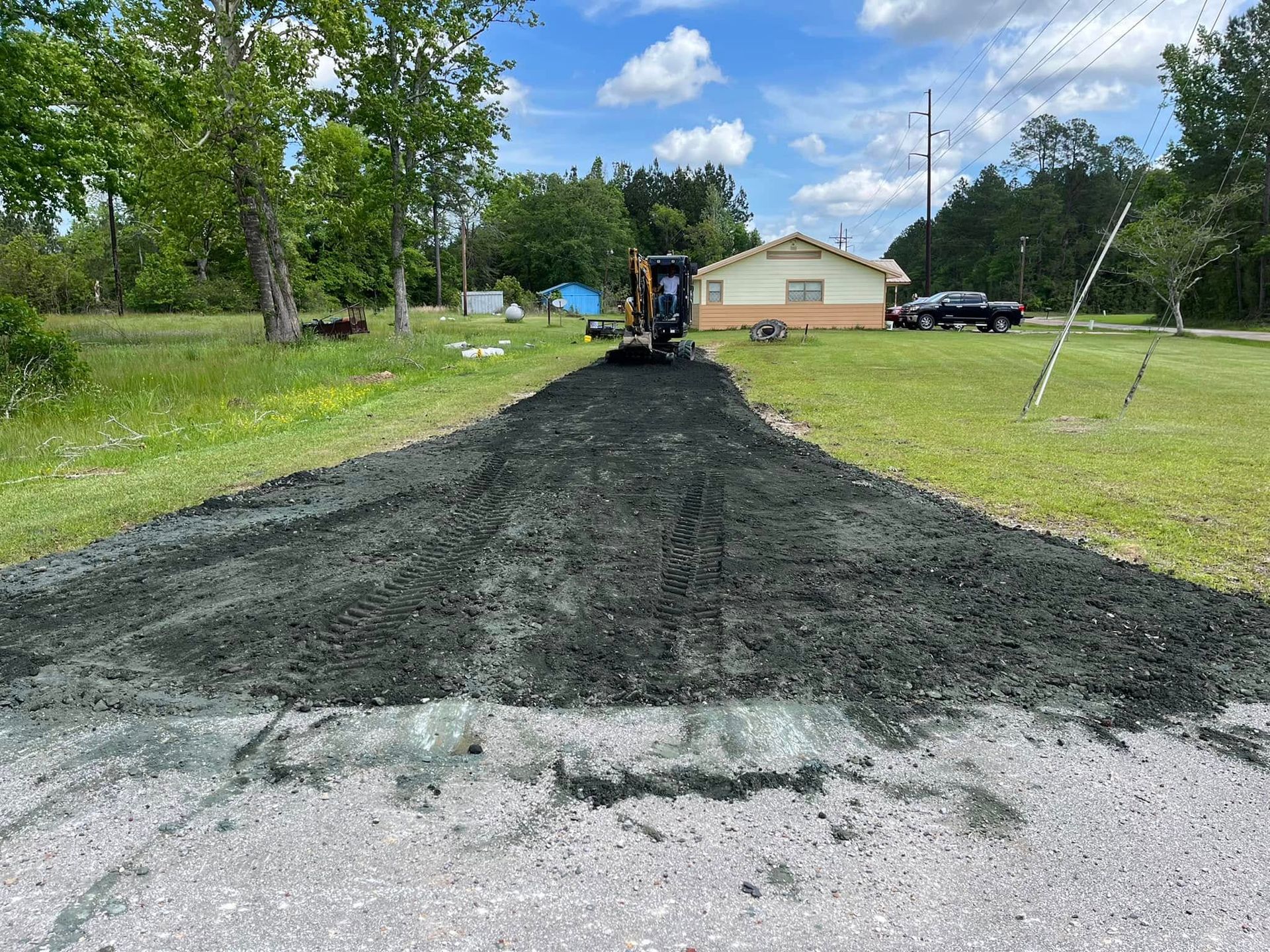 Fresh black gravel driveway being laid toward a beige house with a worker and machinery in the distance