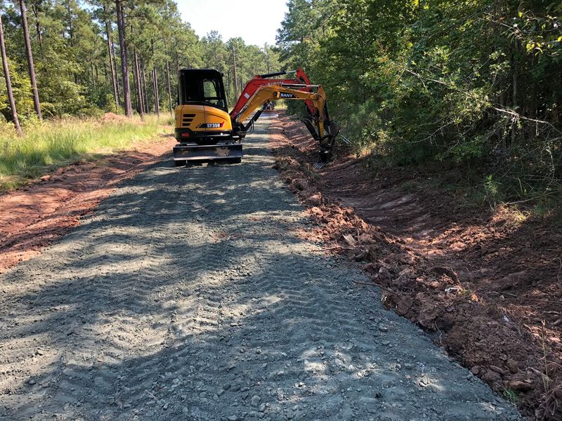 Excavator grading a gravel road beside a wooded path in daylight