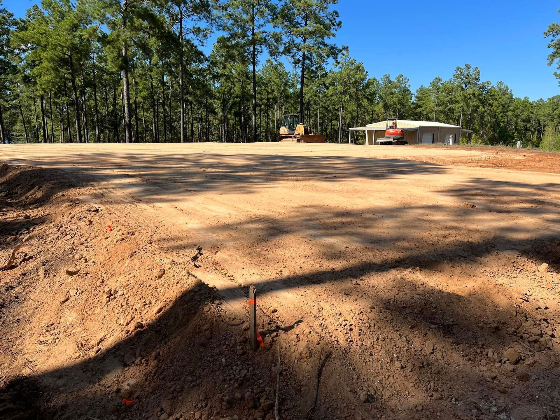 Cleared dirt lot with a red stake in the foreground, trees in back, and a small building under a blue sky.
