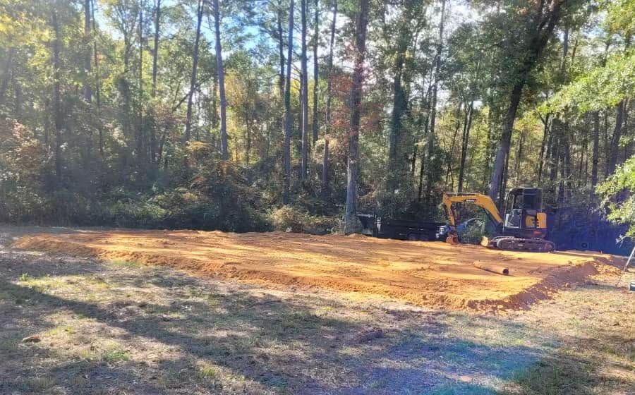 Cleared dirt lot beside trees, with a yellow excavator on the right in sunlight.