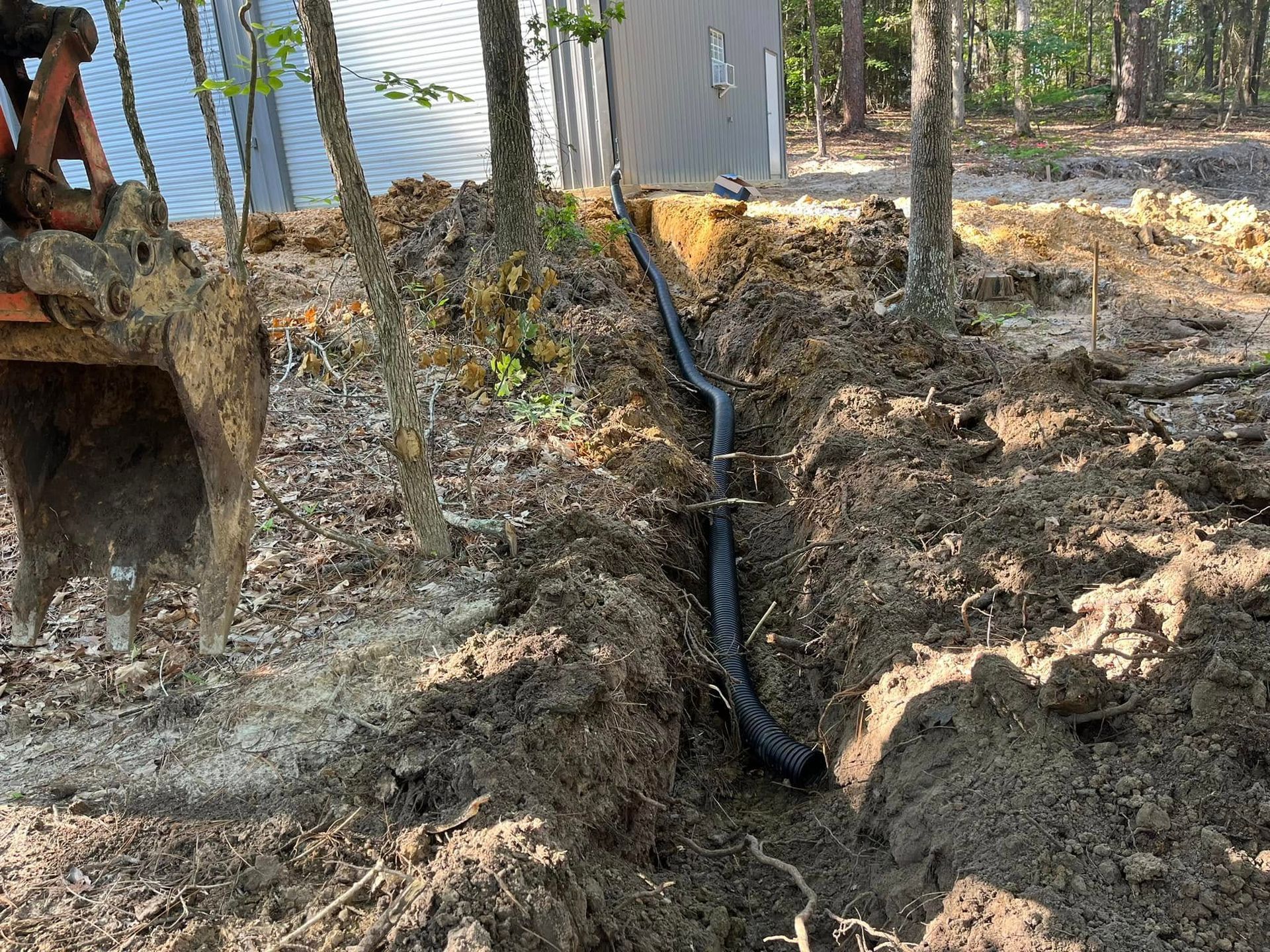 Black pipe runs through a dirt trench beside a house and trees.
