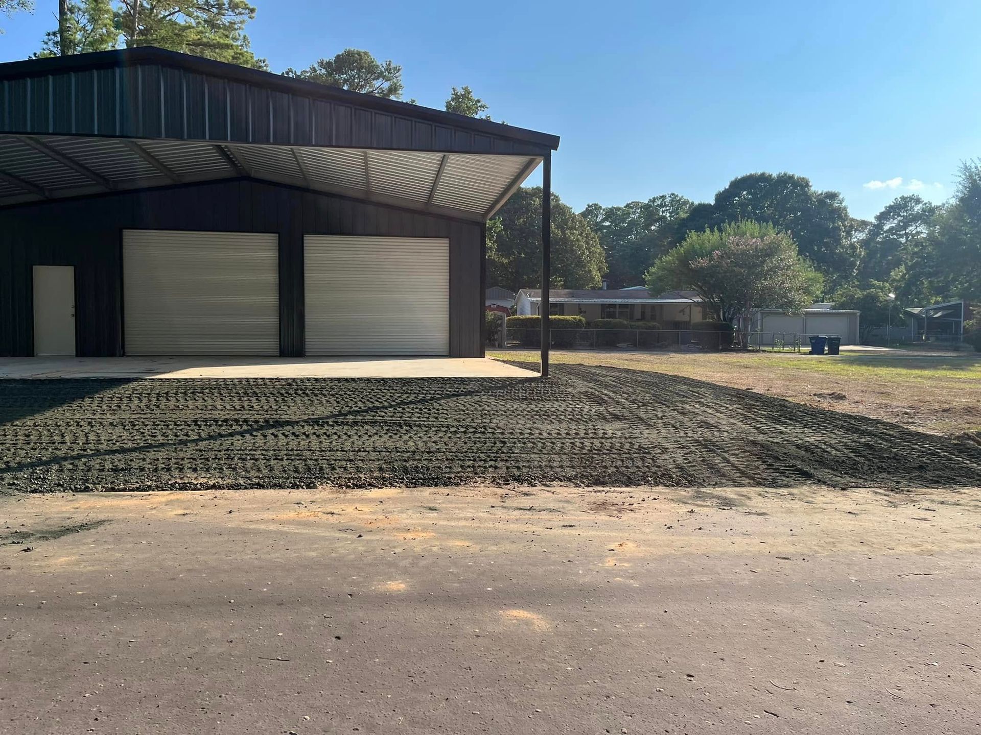 Large metal building with three garage doors beside a sunlit gravel and dirt lot.