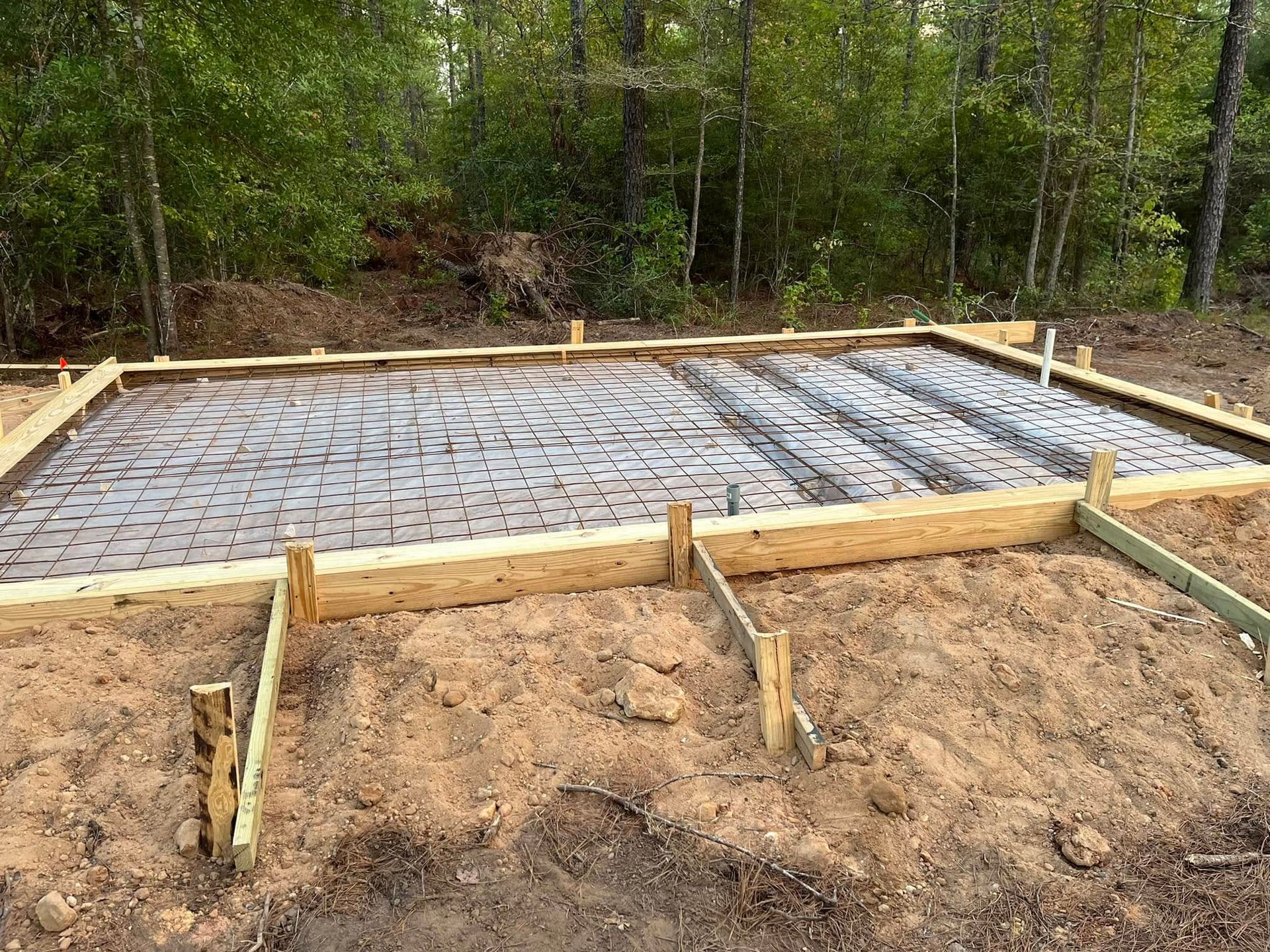 Concrete slab foundation under construction in a wooded area, with wooden forms and rebar grid.