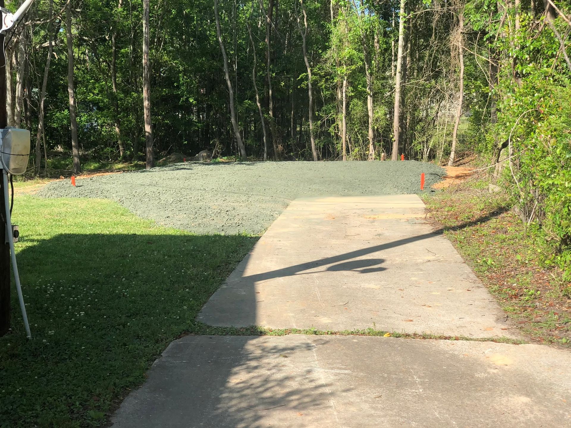 Concrete driveway beside a green gravel pad and wooded area in bright sunlight