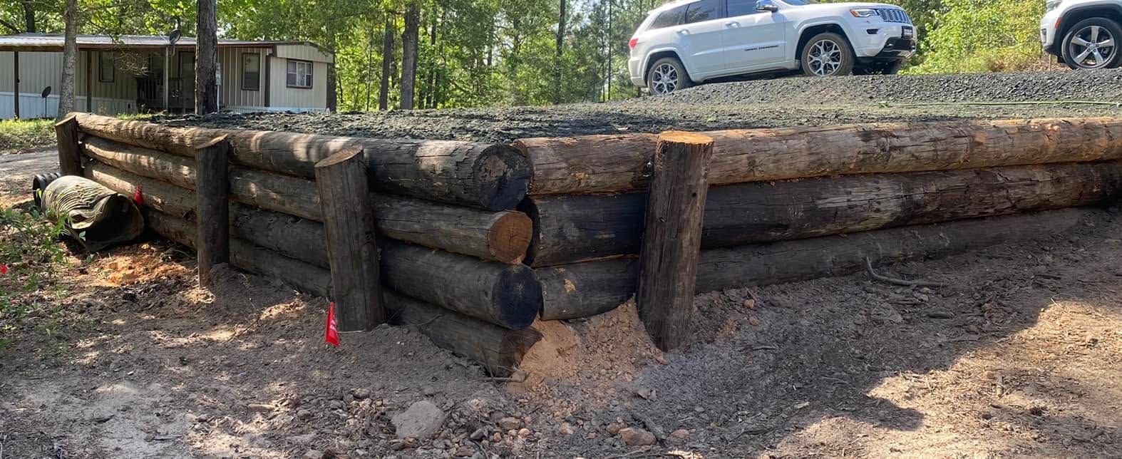 Log cabin-style retaining wall made of stacked logs on a gravel lot, with a white SUV in the background.