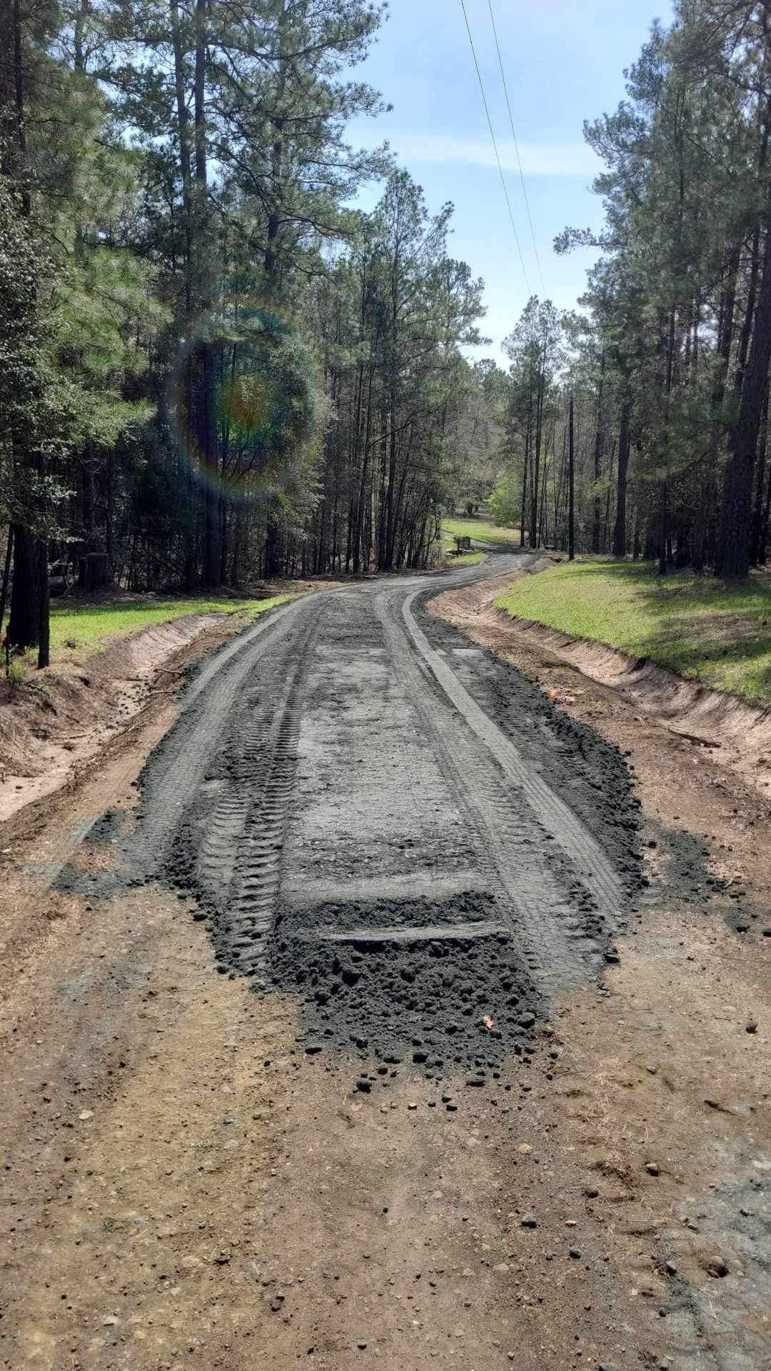 Gravel road under construction through a pine forest, with fresh dirt shoulders and a narrow paved lane.