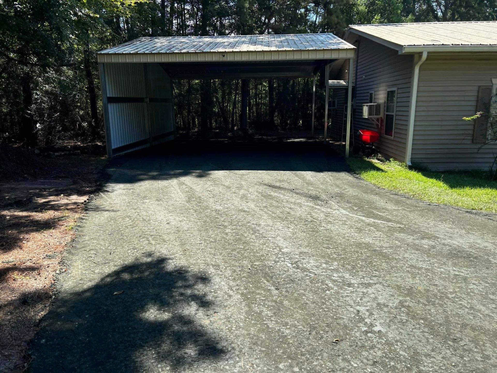 Gravel driveway leading to a covered carport beside a house, with trees and bright sunlight.