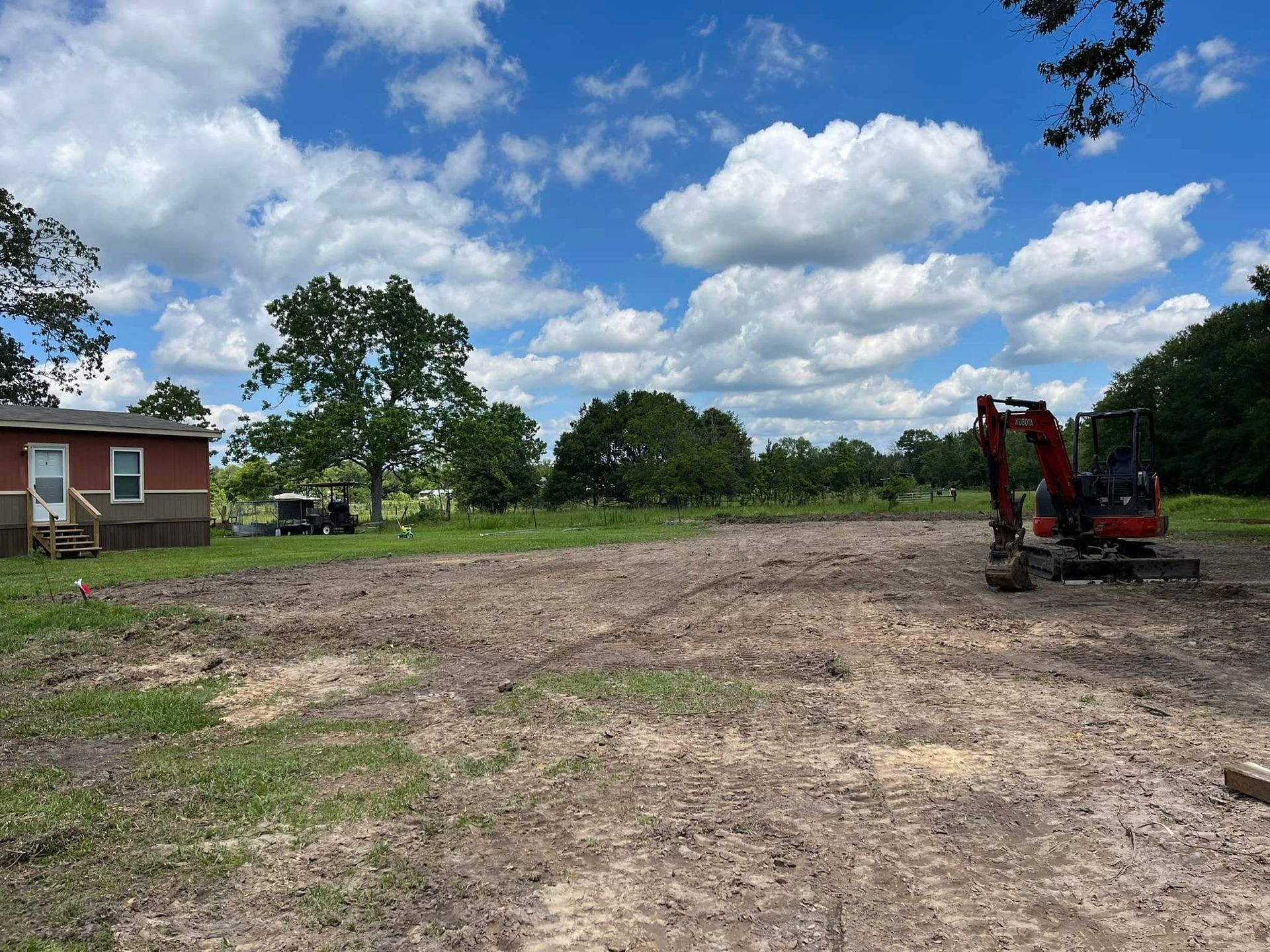 Cleared dirt lot with a red excavator under a blue sky, beside a small house and trees