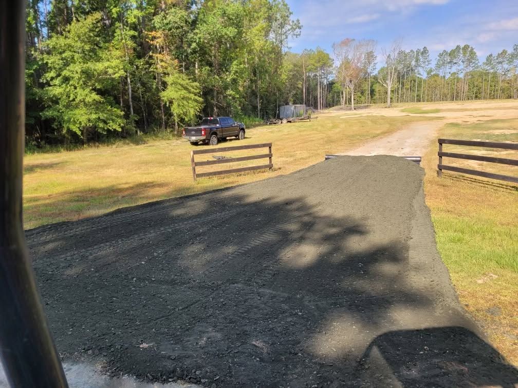 Gravel driveway entrance with wooden gates, a parked dark truck, and trees along a sunny rural road