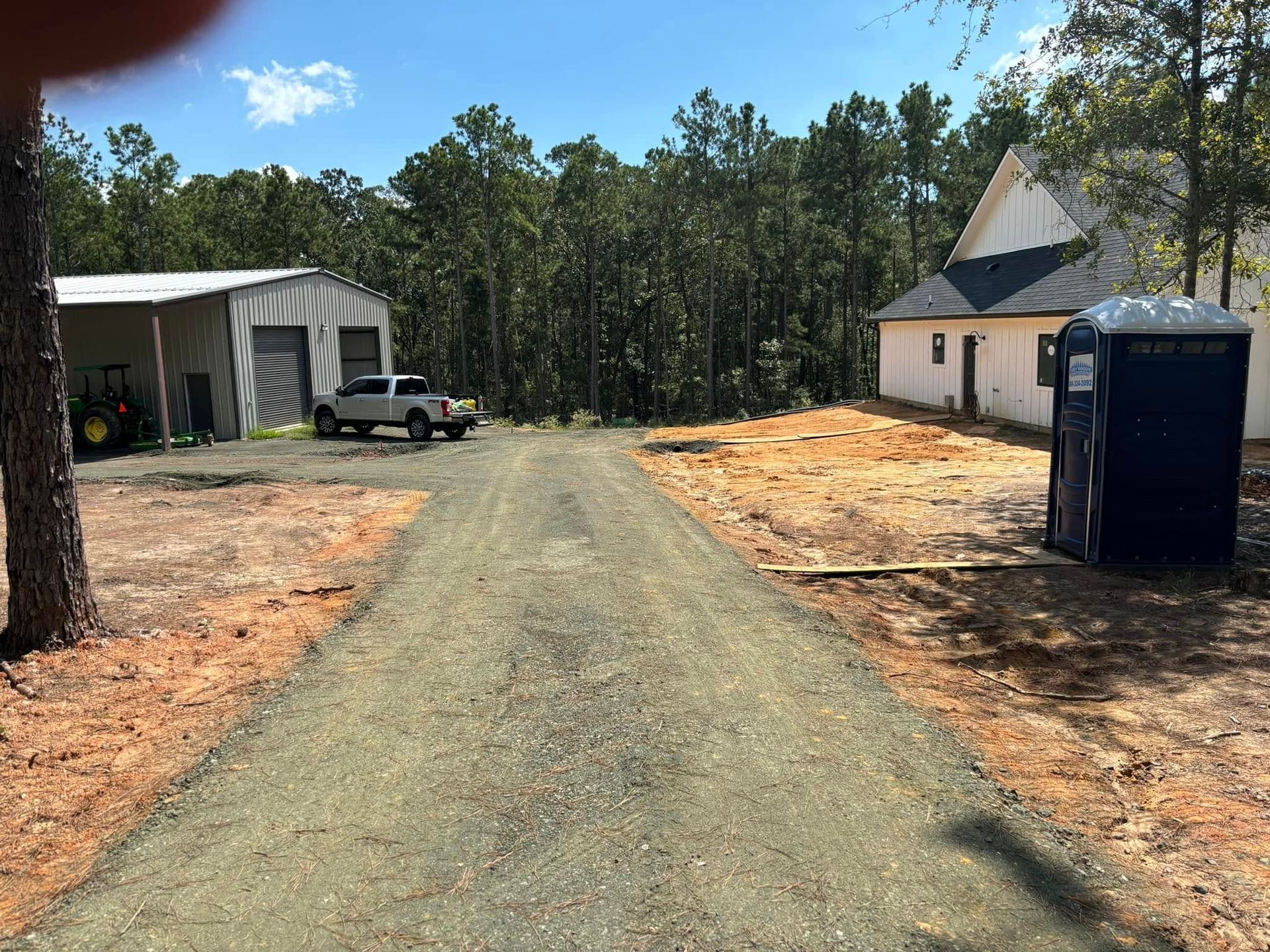 Gravel driveway between a garage and house, with a portable toilet on the right and trees in back.