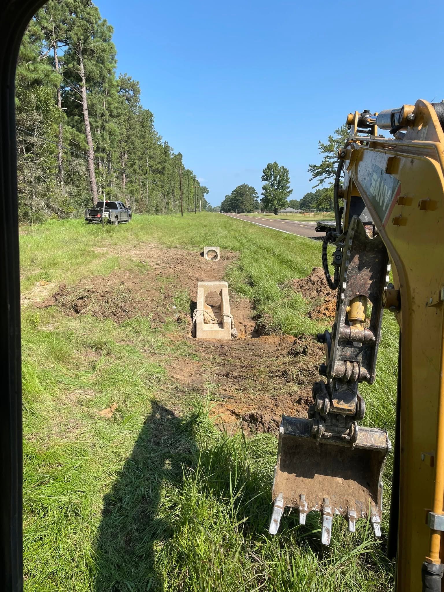 Yellow excavator working beside a grassy roadside ditch under a clear blue sky