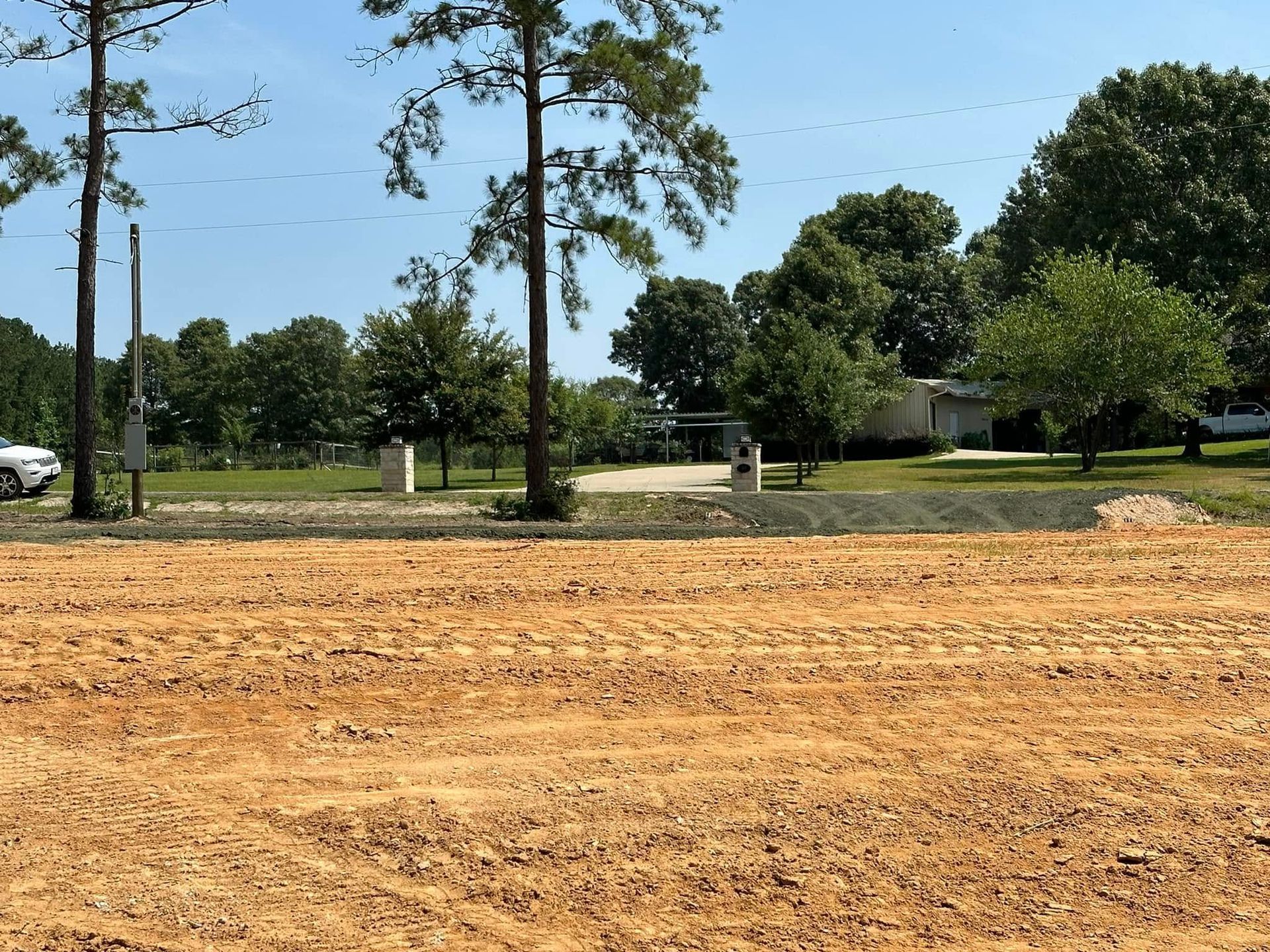 Freshly graded dirt lot with trees and houses in the background under a clear sky