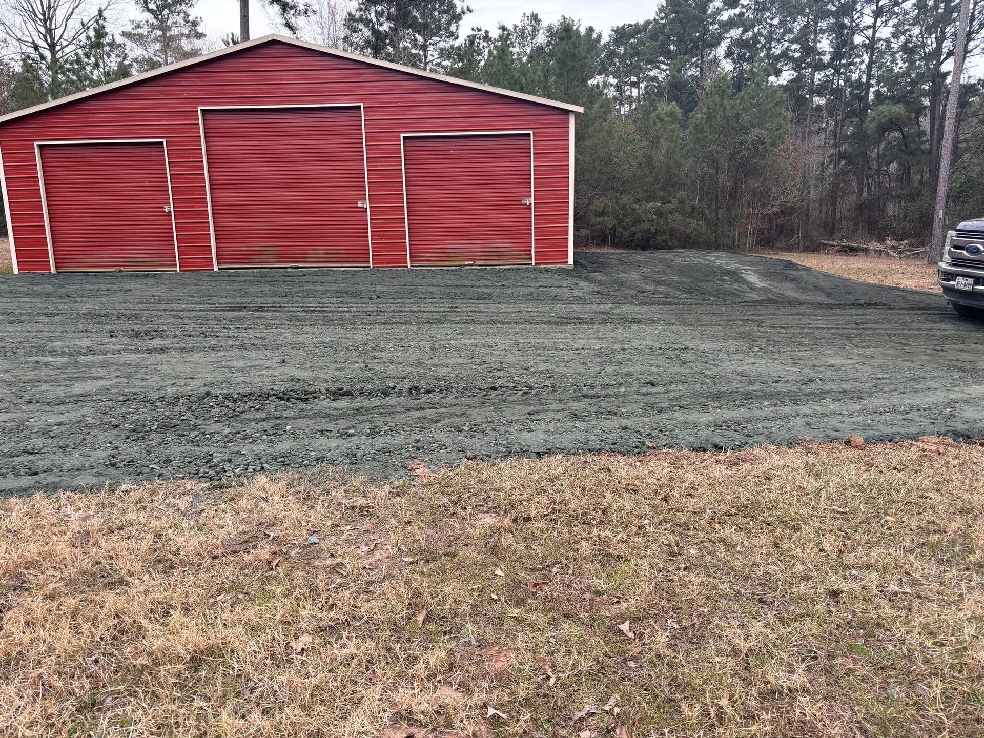 Red barn with white trim on a cleared gravel lot beside a wooded area