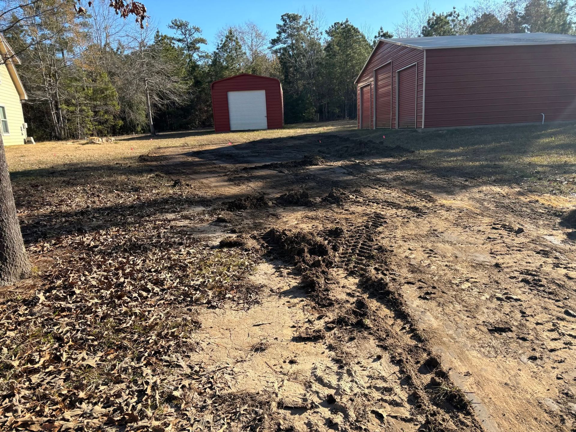 Dirt yard with ruts and bare patches, beside two red sheds under a blue sky.
