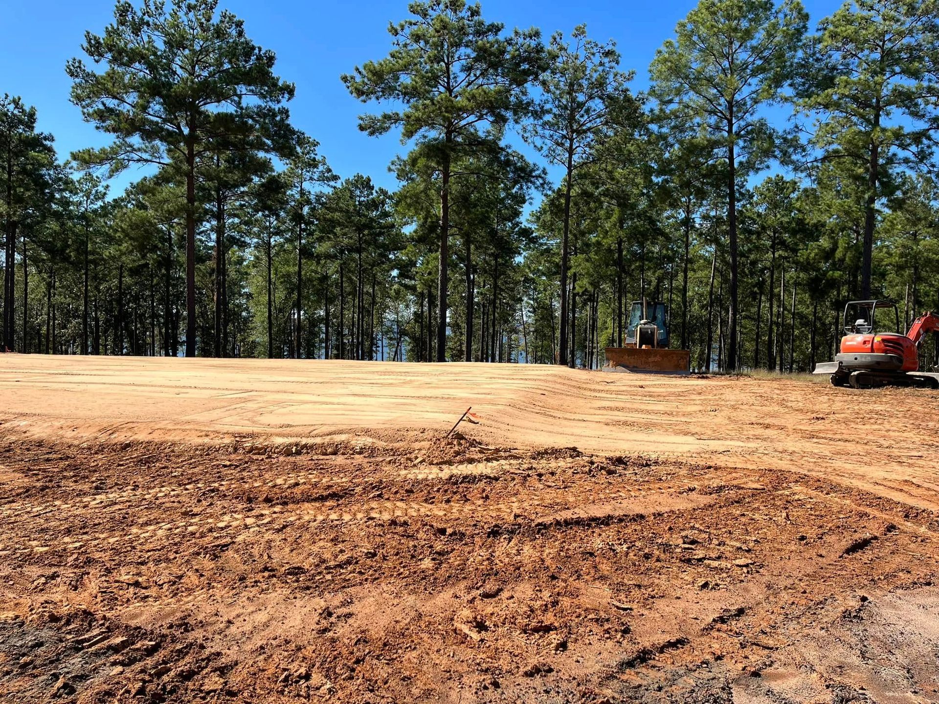 Cleared dirt lot with pine trees in the background and a red excavator at the right edge
