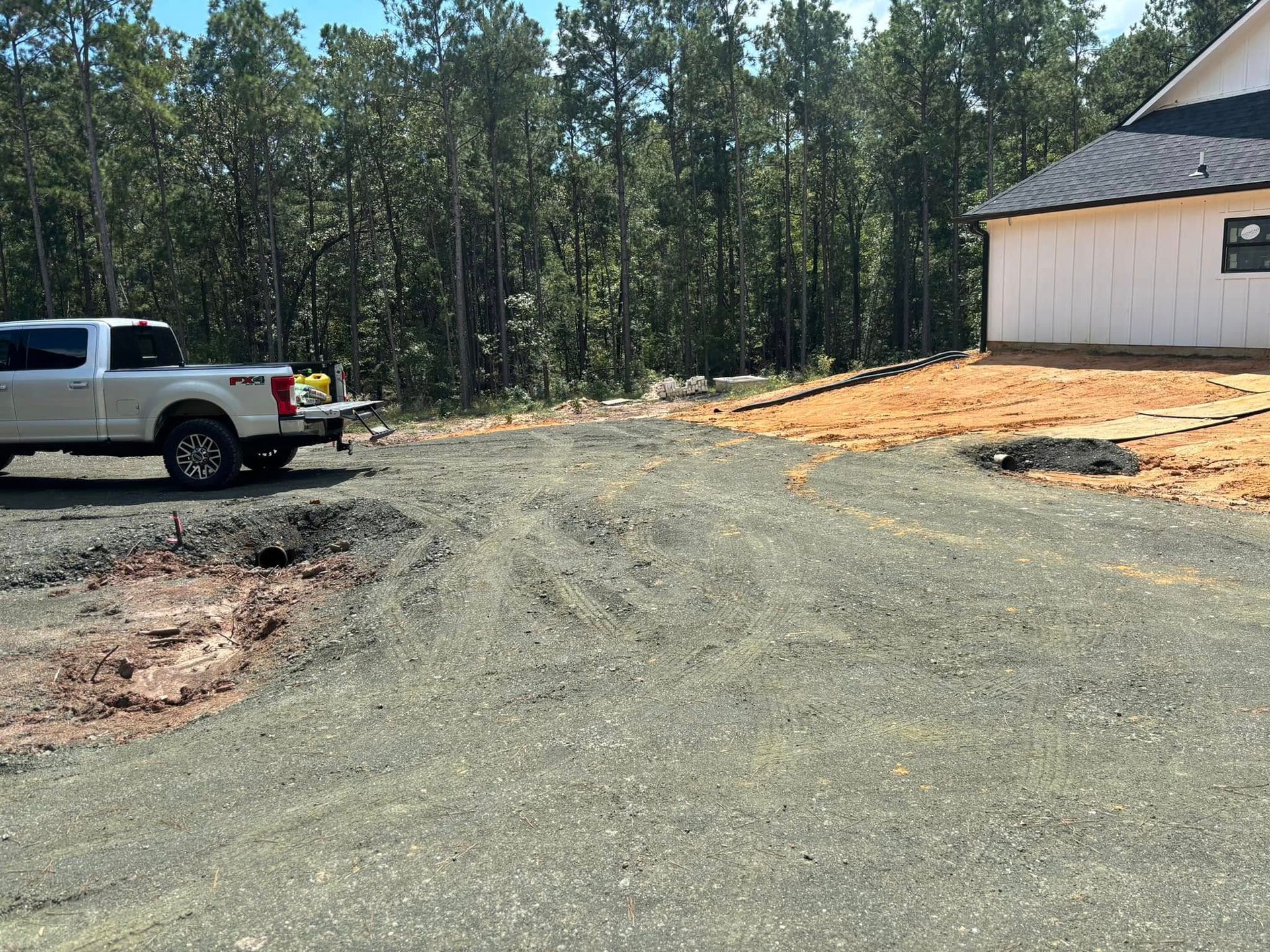 Gravel driveway under construction beside a white house, with a pickup truck and wooded background.