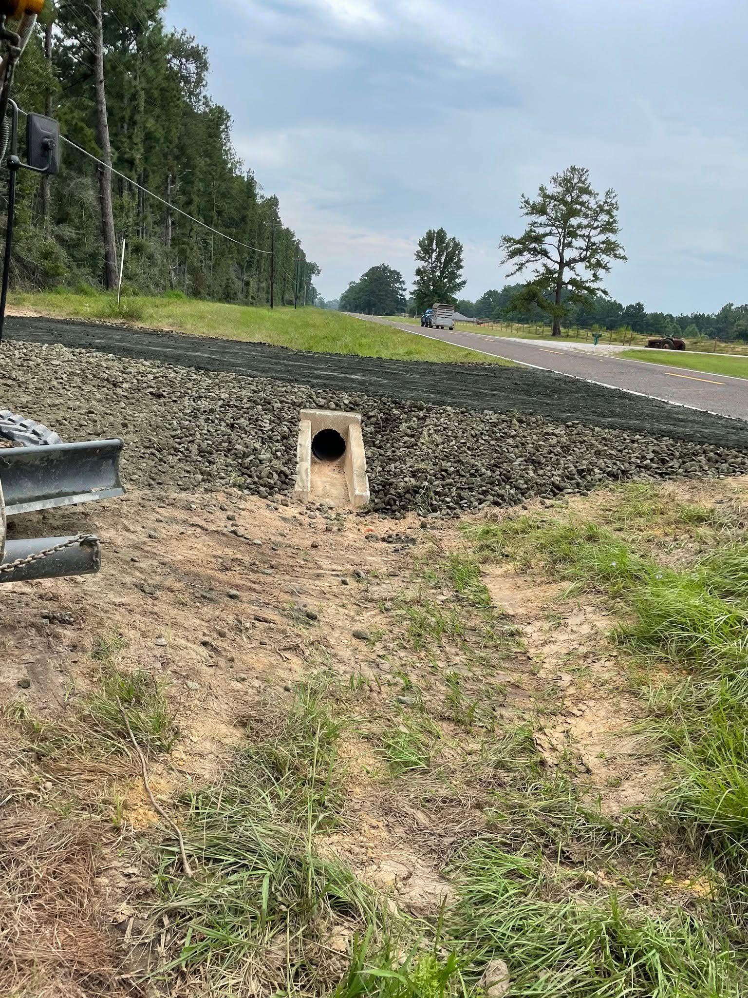 Roadside culvert opening in a gravel embankment beside a paved road and trees