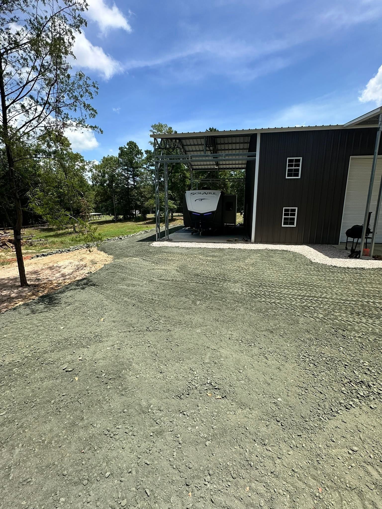 Gravel driveway leading to a black building and carport under a blue sky, with trees and landscaping nearby