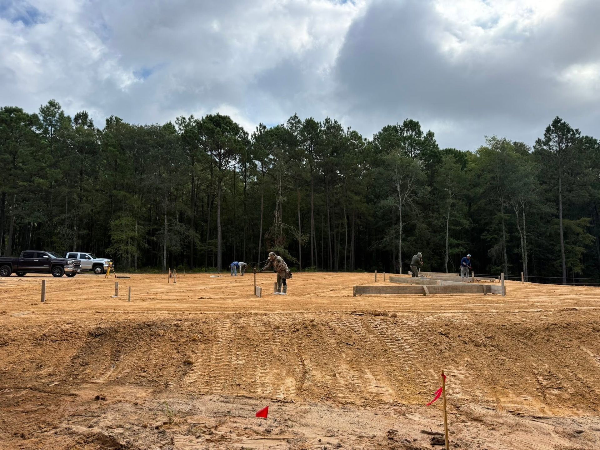 Construction site with graded dirt lot, a worker at center, red flags, trees, and cloudy sky in the background