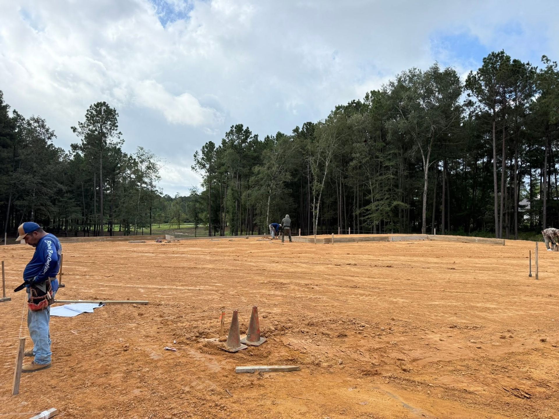 Workers surveying a cleared dirt lot with trees in the background under a cloudy sky