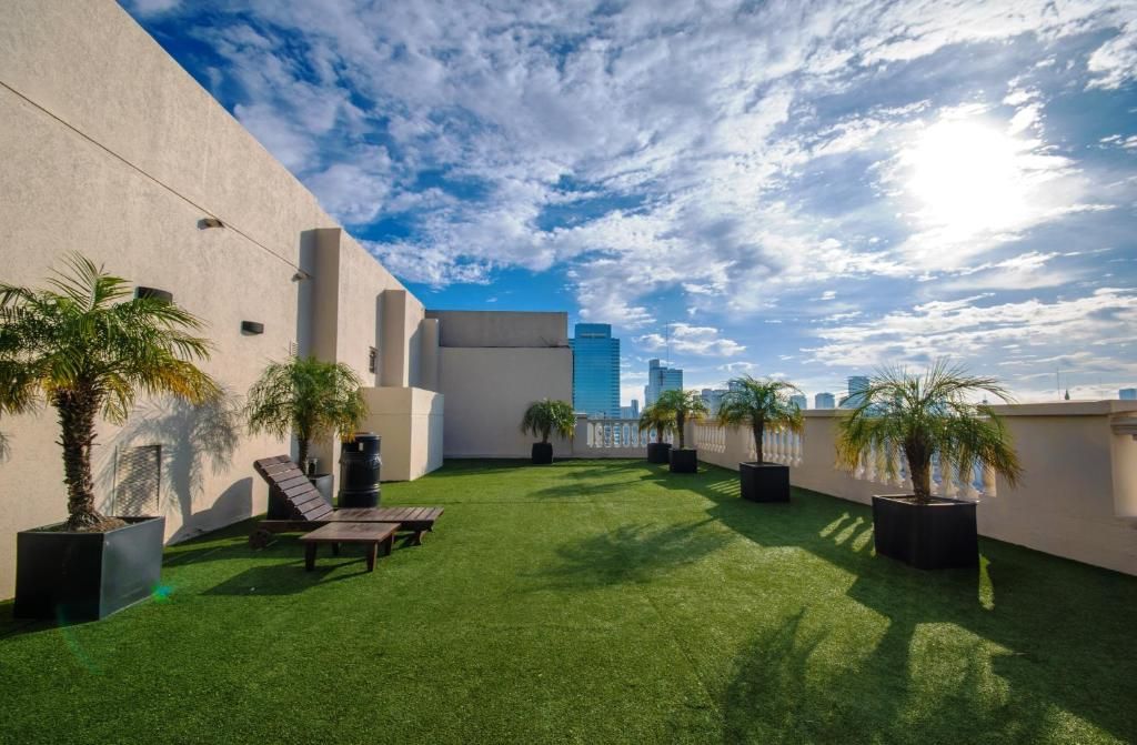 Una terraza en la azotea con césped artificial, palmeras en macetas y tumbonas, con vistas al soleado horizonte de la ciudad.