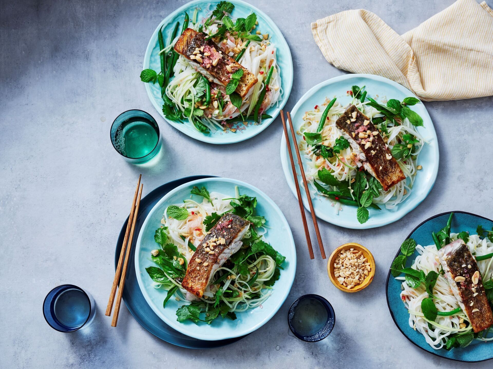 Four plates of seared salmon fillets served over rice noodles, herbs, and greens on a light grey textured background.
