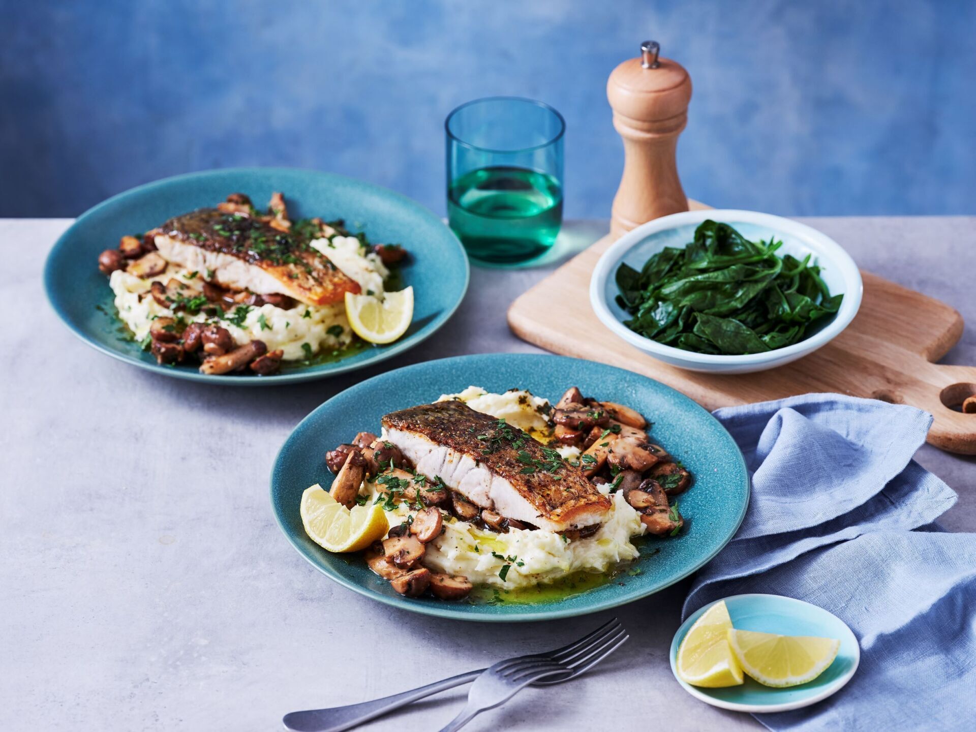 Two plates of pan-seared fish on creamy mashed potatoes with mushrooms and lemon wedges, served with a side of spinach.