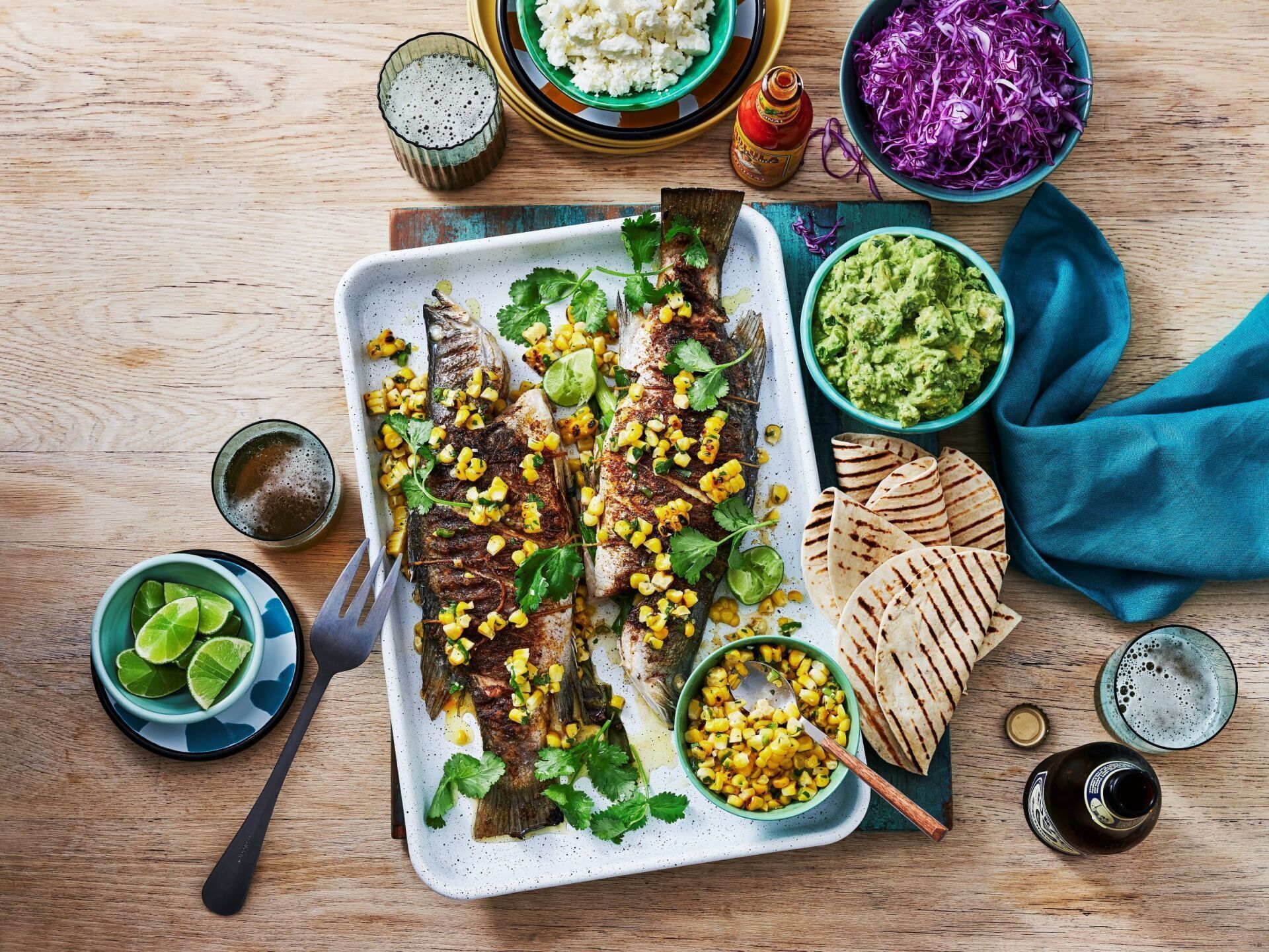 A white tray with two grilled fish topped with corn and cilantro, surrounded by bowls of sides, tortillas, and a drink.