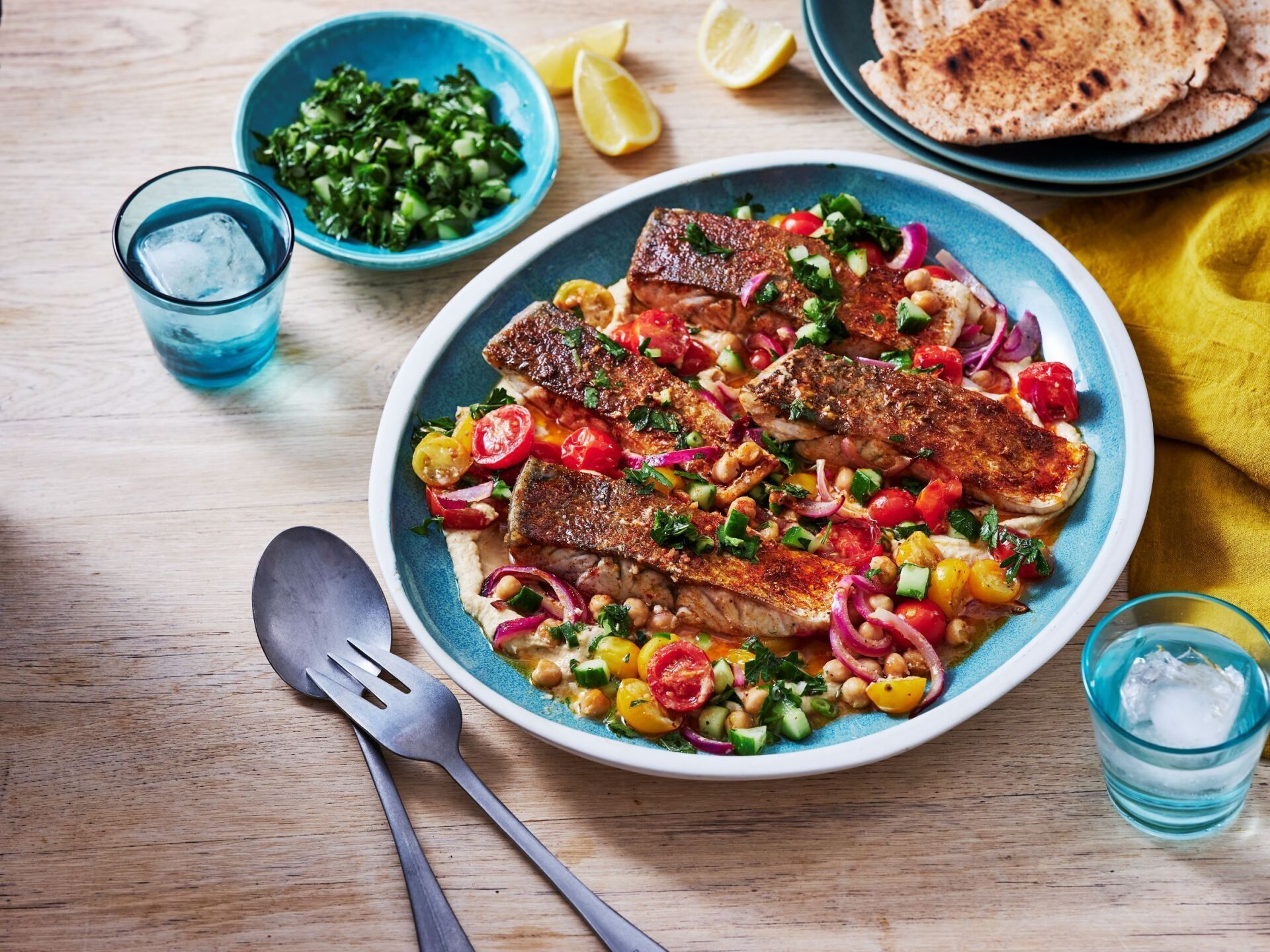A plate of seared fish fillets served over chickpeas and a salad of red onions and tomatoes on a light wooden table.