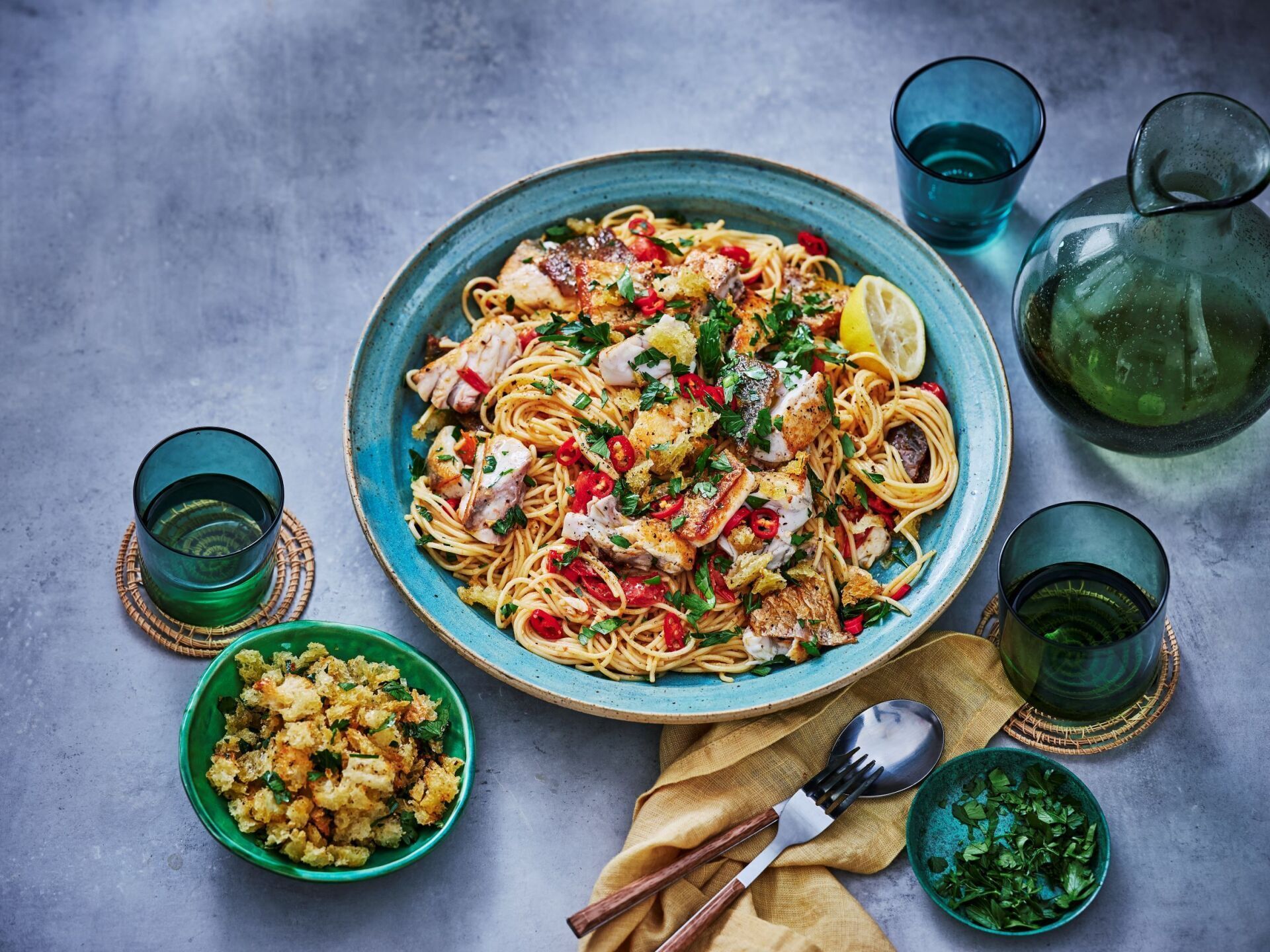 A large blue bowl of pasta with white fish, herbs, and red chili on a gray table with drinks and side dishes.
